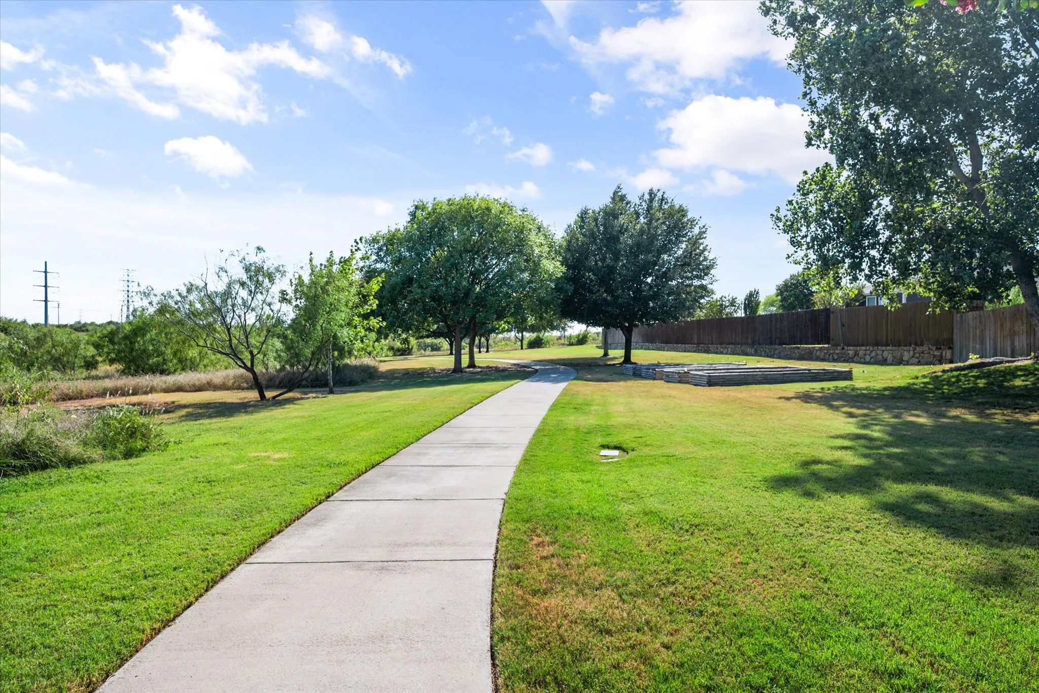 Expansive green space featuring a concrete pathway, mature trees, a wooden fence, and stone-accented landscaping beds