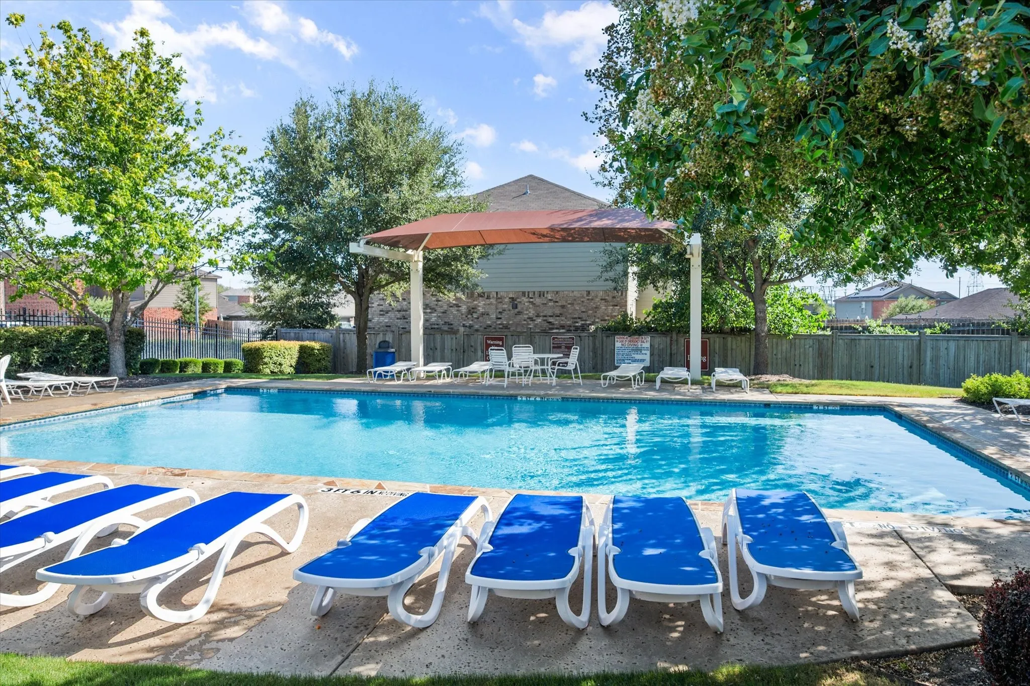 Community swimming pool with concrete decking, surrounding green foliage, and a shade structure over a patio area