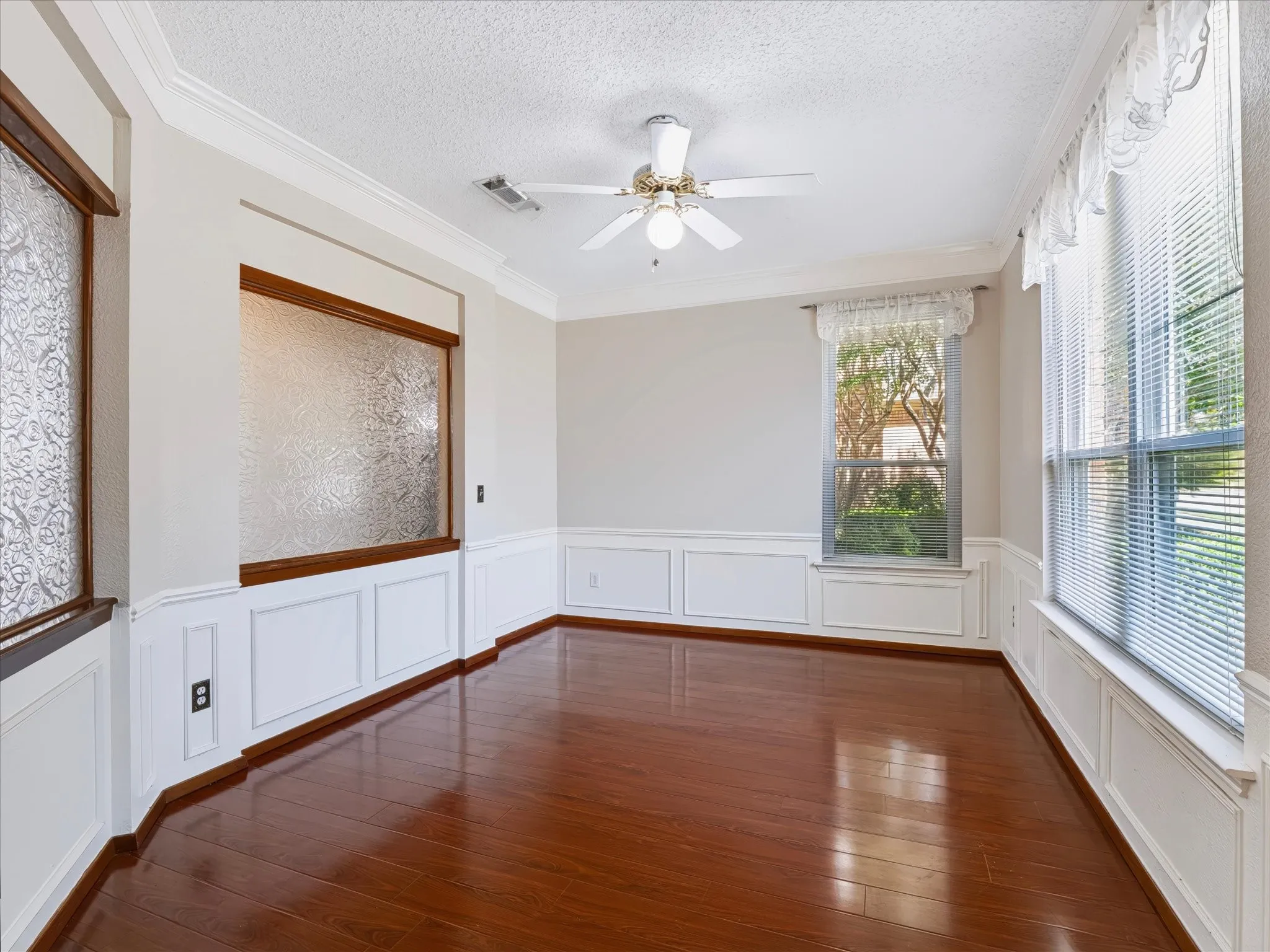 Rich wood-finish flooring, white wainscoting, crown molding, and a ceiling fan with integrated lighting