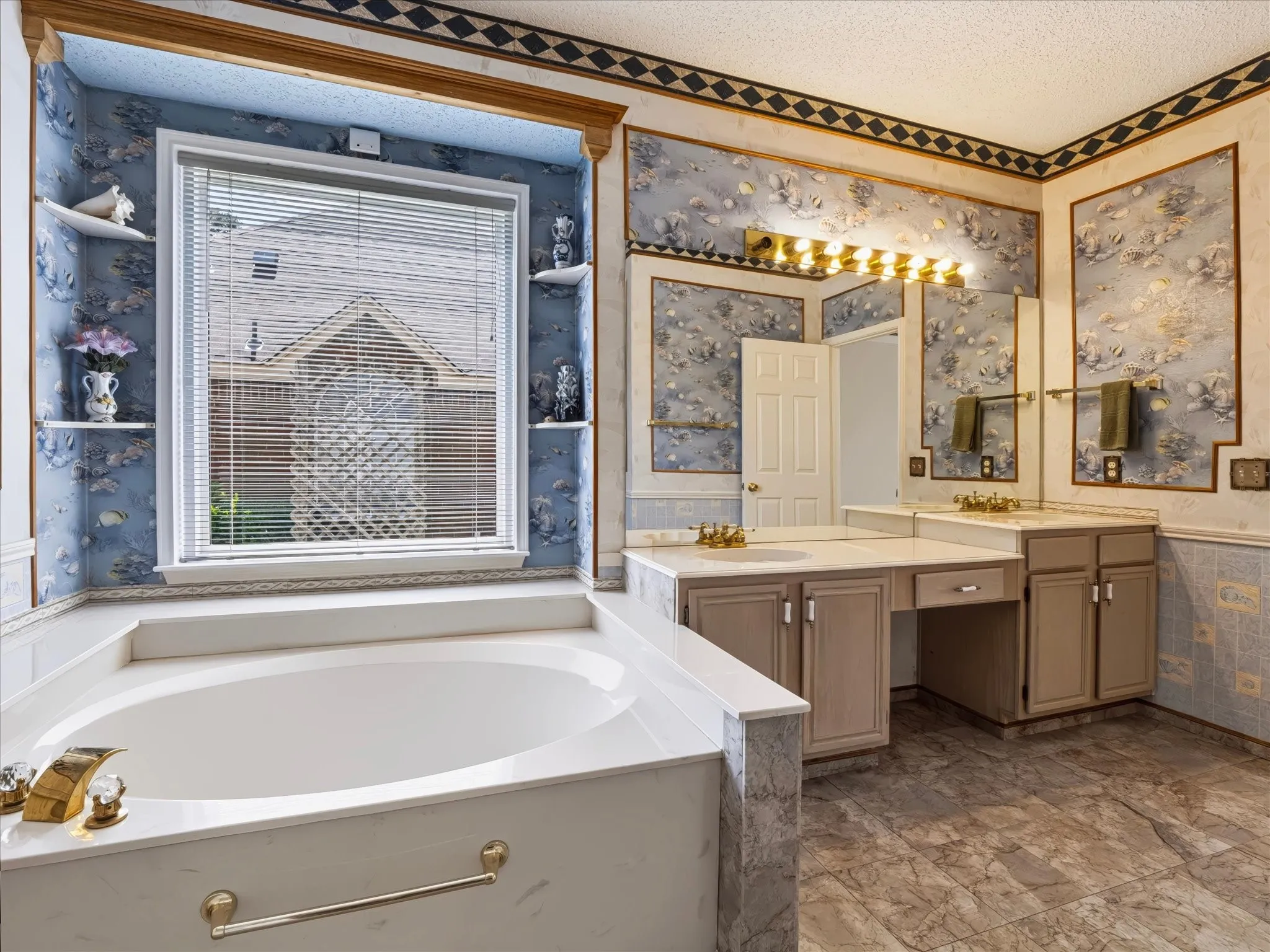 Spa-inspired primary bathroom featuring a soaking tub with brass hardware, dual-sink vanity with wood-finish cabinetry and warm-toned countertops, large illuminated mirrors, and decorative wallpaper