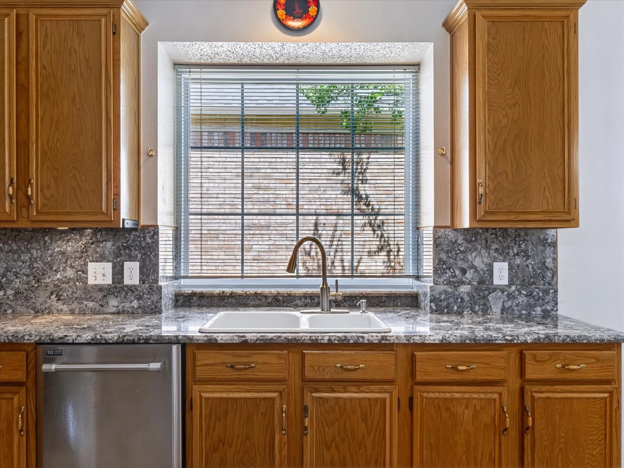 Kitchen featuring wood cabinetry, a double basin sink with a gooseneck faucet, a stainless steel dishwasher, and stone countertops with a matching backsplash