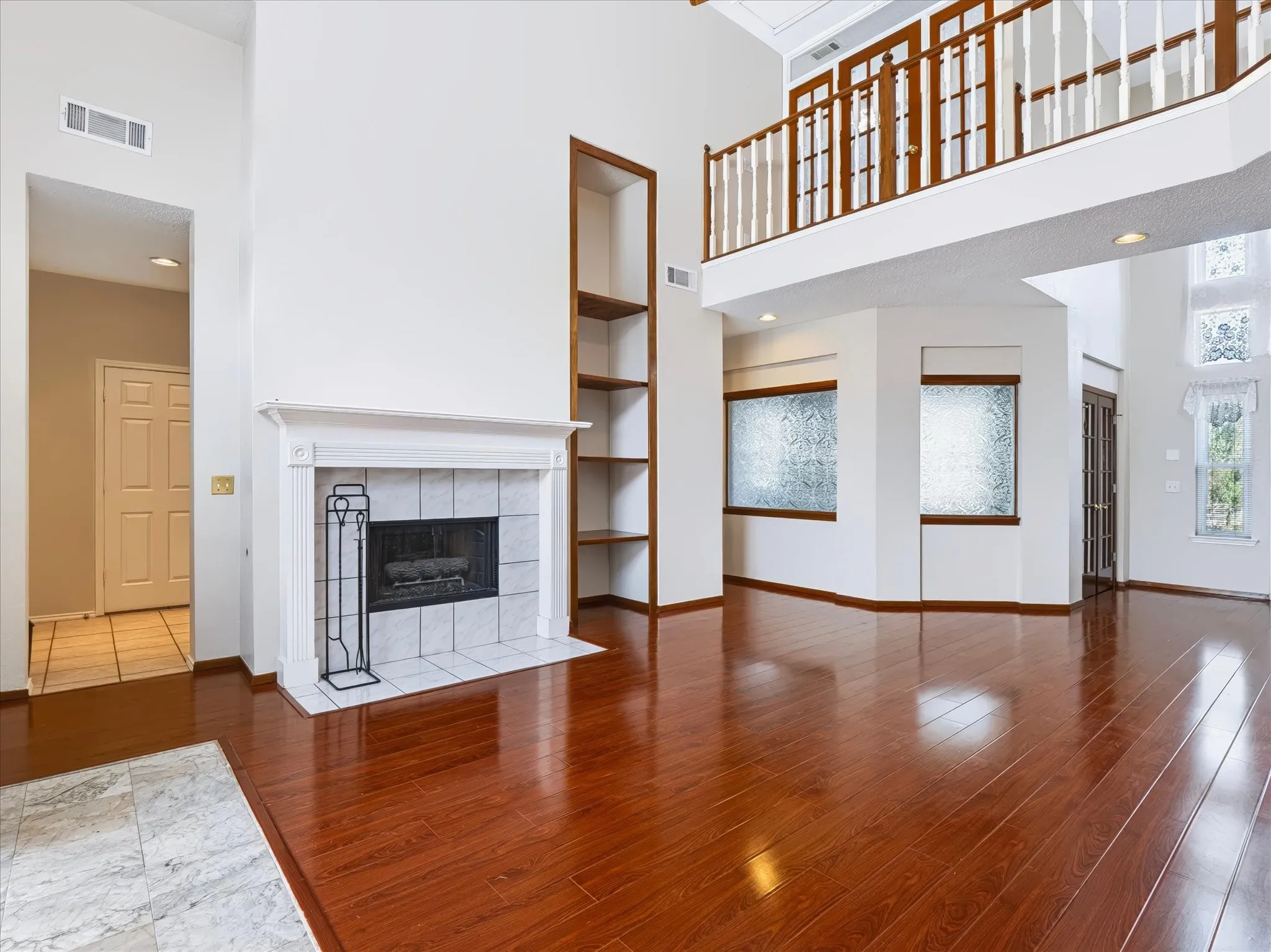 Hardwood-finish flooring, a white mantel fireplace with tile surround, built-in shelving, and a second-story interior balcony with wood railing
