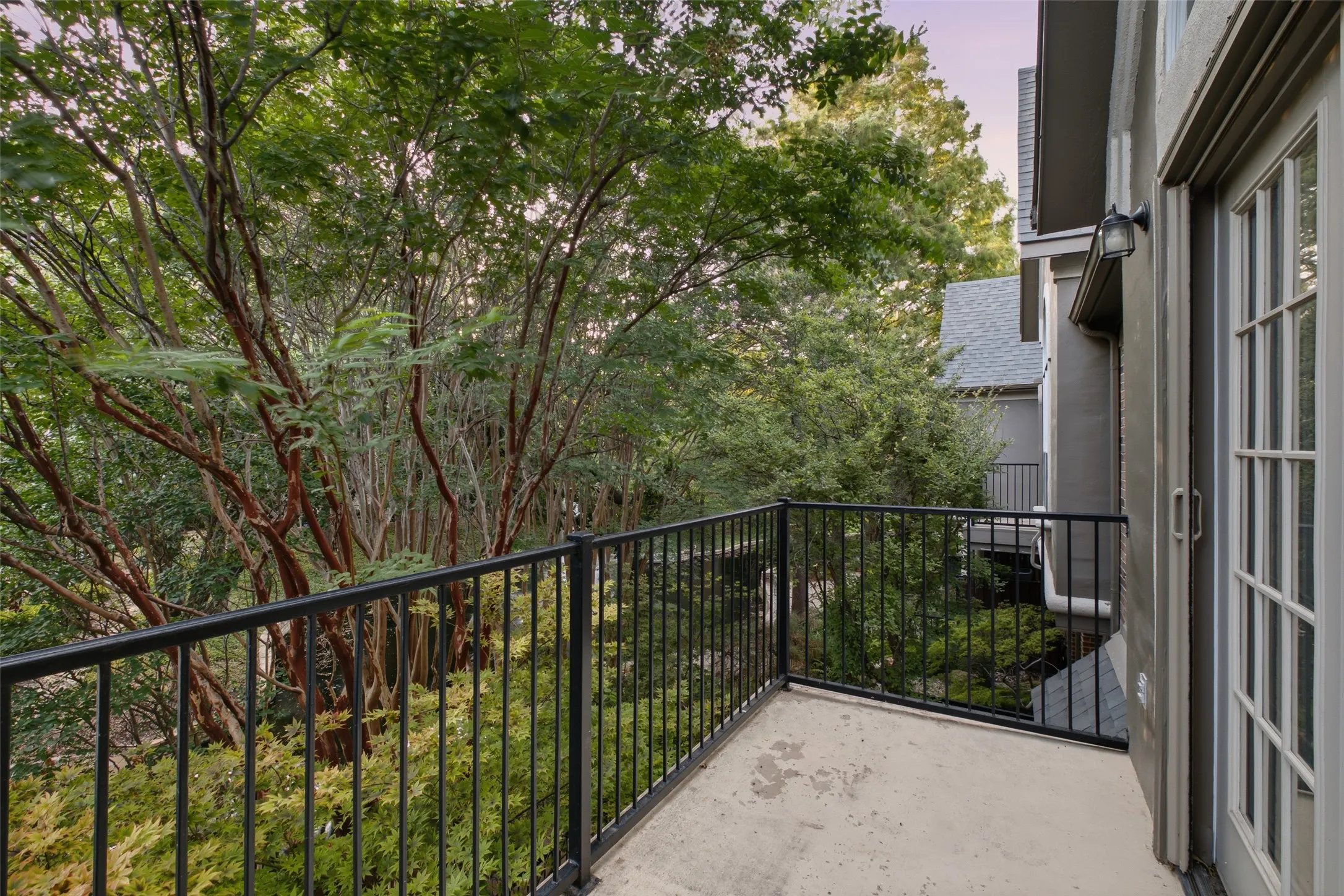 Balcony with black metal railings, concrete flooring, and an exterior wall sconce, featuring mature trees and neighboring structures