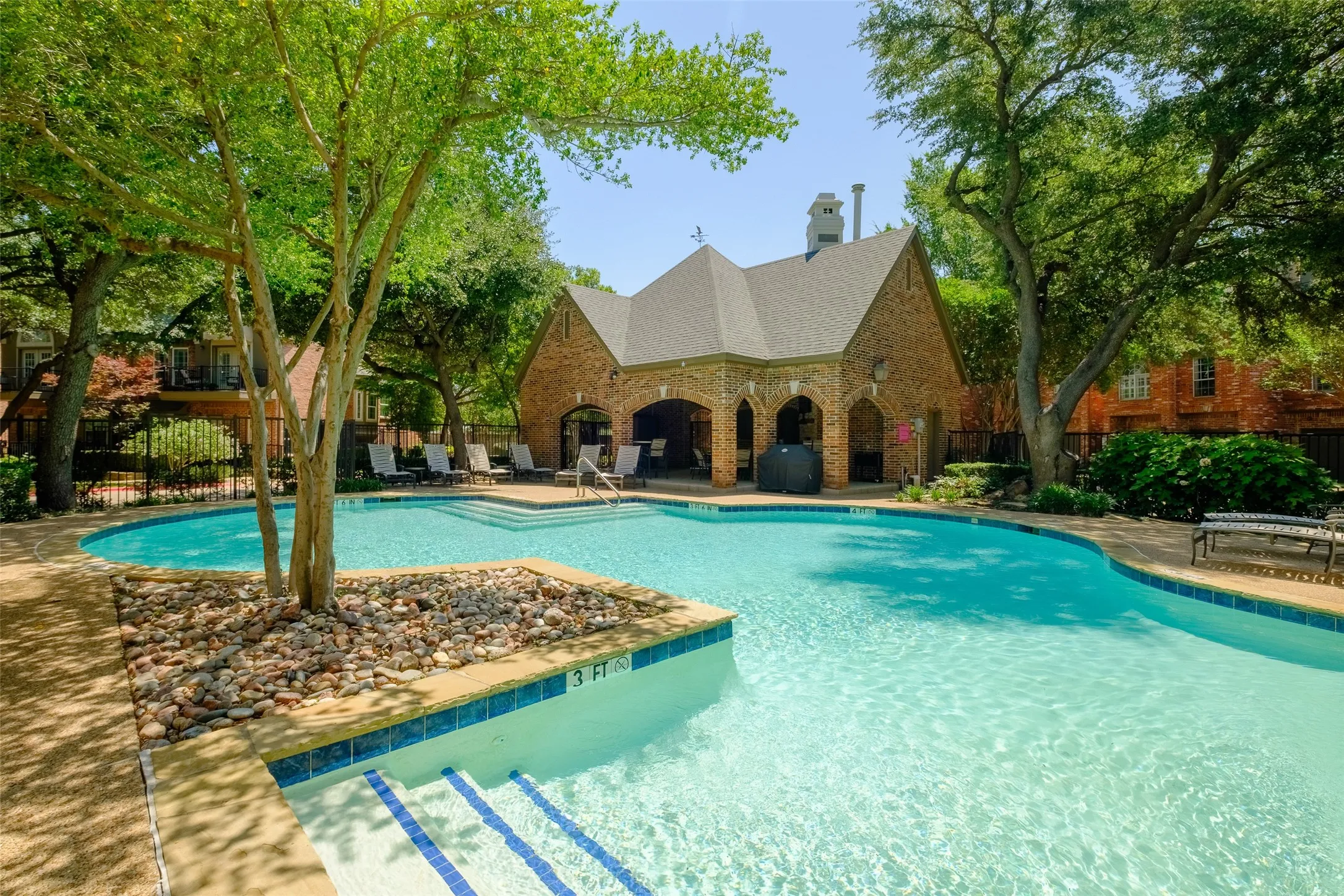 Expansive swimming pool with integrated steps, surrounded by mature trees and a brick-built clubhouse featuring arched openings