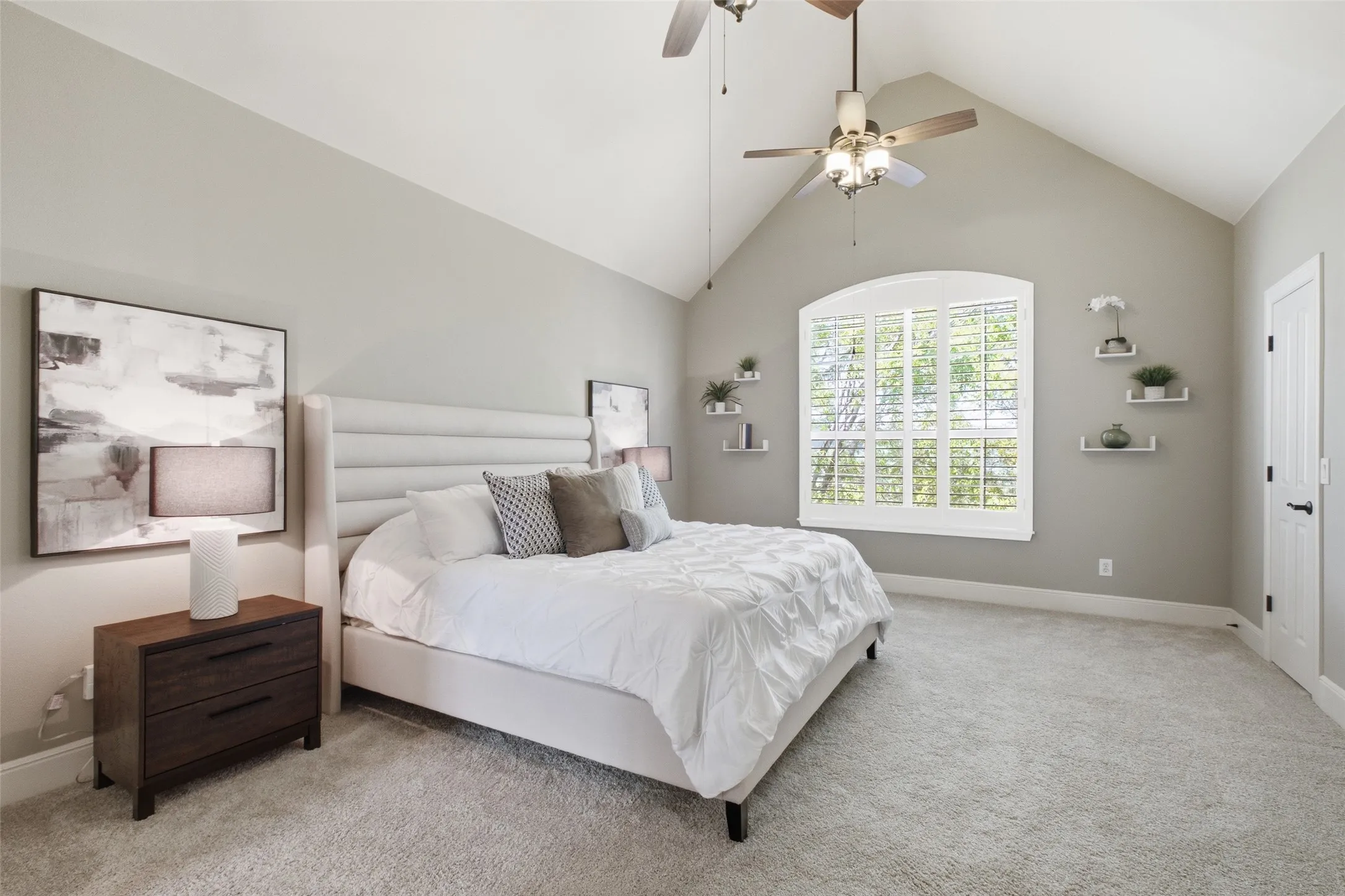 Vaulted ceiling with dual ceiling fans, arched window with plantation shutters, light gray wall tones, and plush carpeting