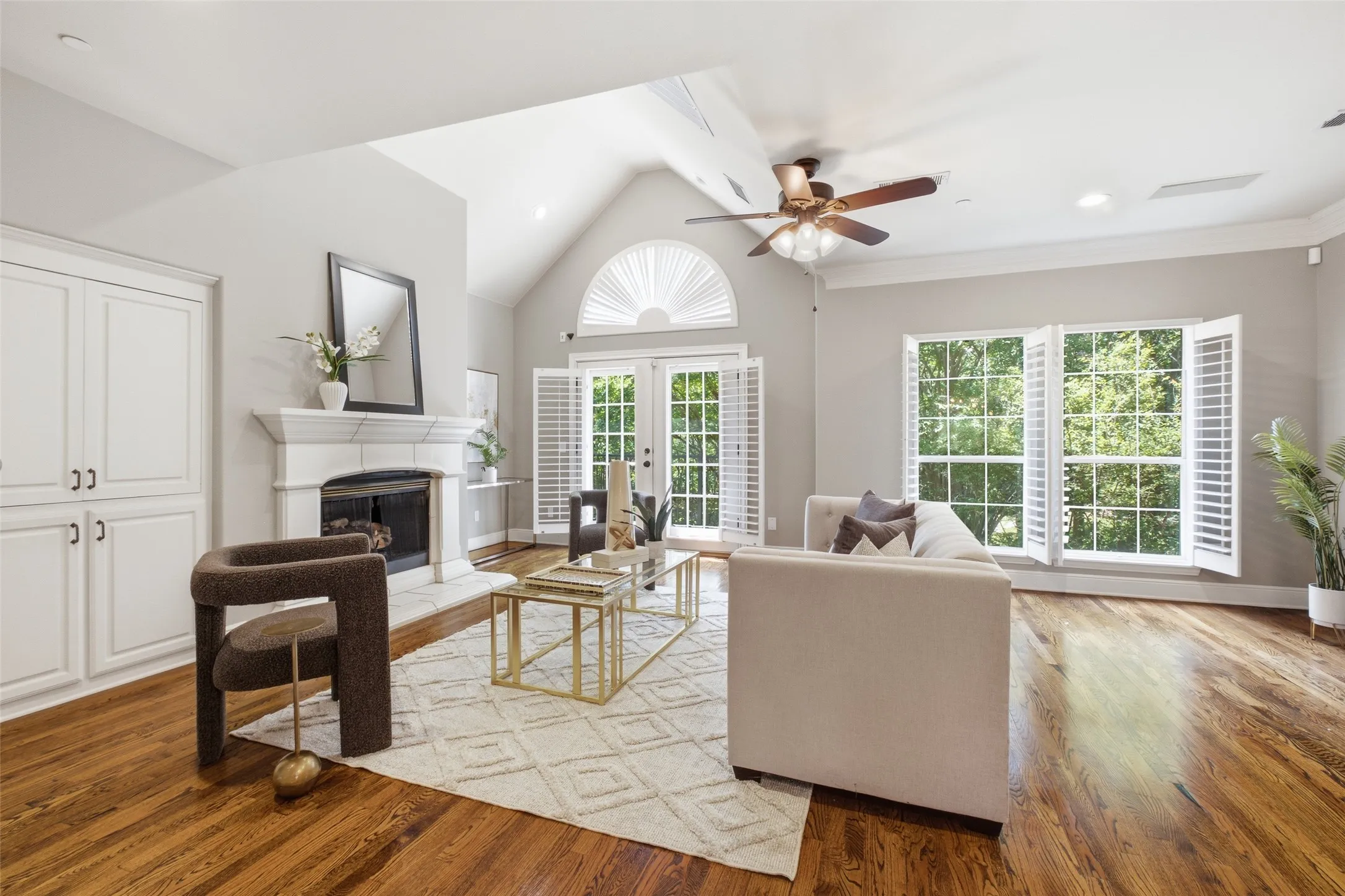 Vaulted ceiling with a ceiling fan, fireplace with a mantel, built-in cabinetry, wood-finish flooring, and multiple windows with plantation shutters