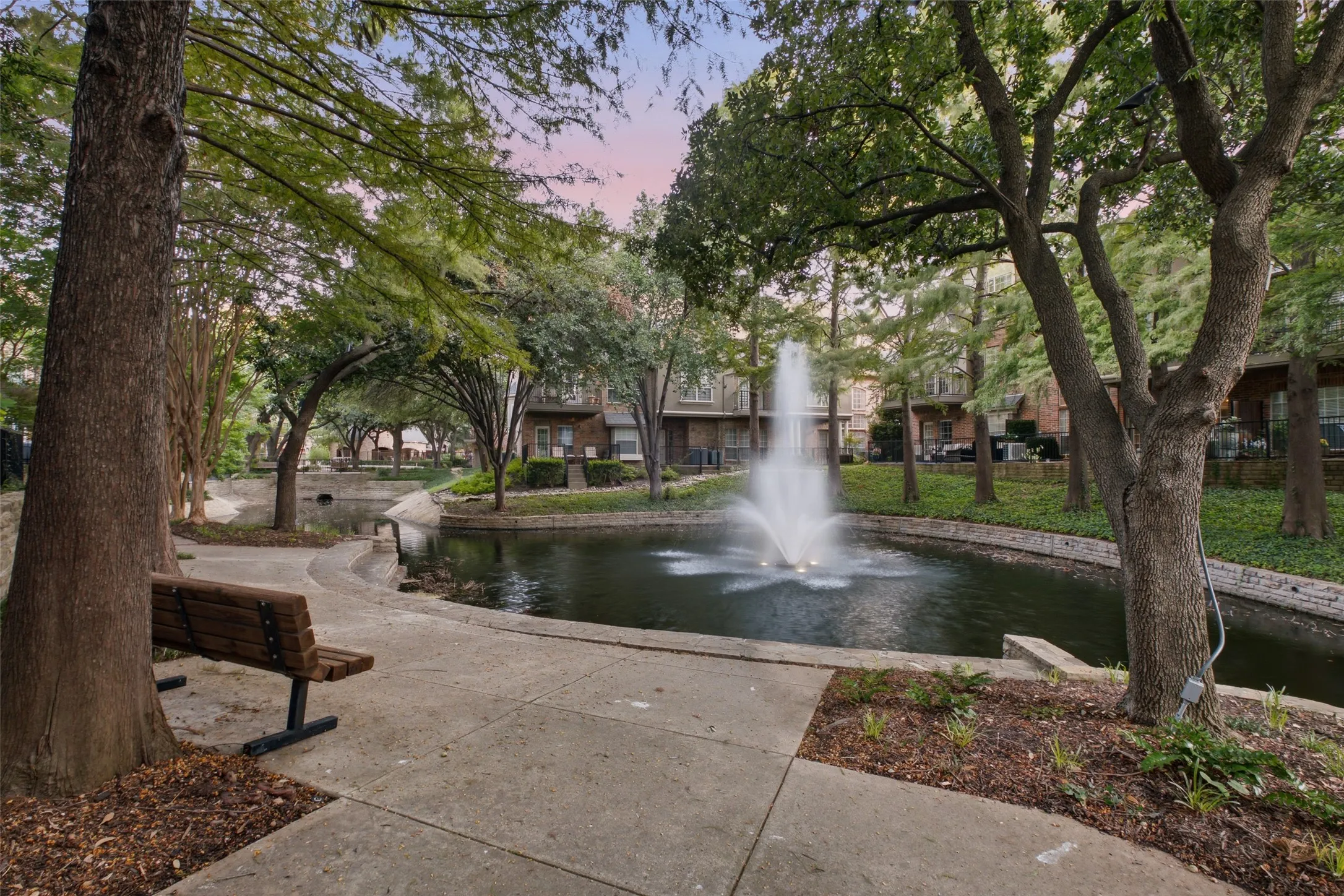 Landscaped grounds featuring mature trees, a tranquil pond with a fountain, and concrete pathways