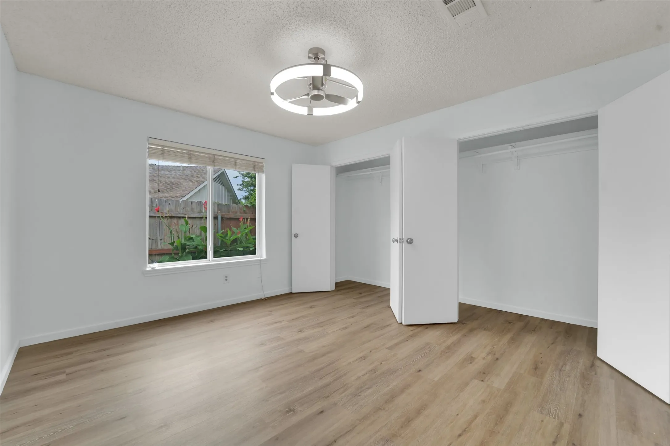 Bright, spacious bedroom featuring wood-finish flooring, abundant natural light from a large window, dual closets with white doors, and a modern recessed ceiling light fixture