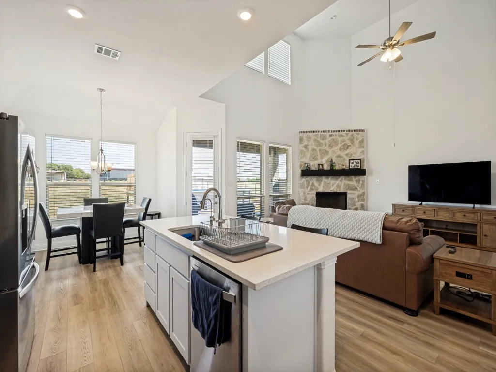 Open concept living area featuring wood-finish flooring, vaulted ceilings, a stone-faced fireplace with a dark mantel, white shaker cabinetry, and a kitchen island with a double basin sink