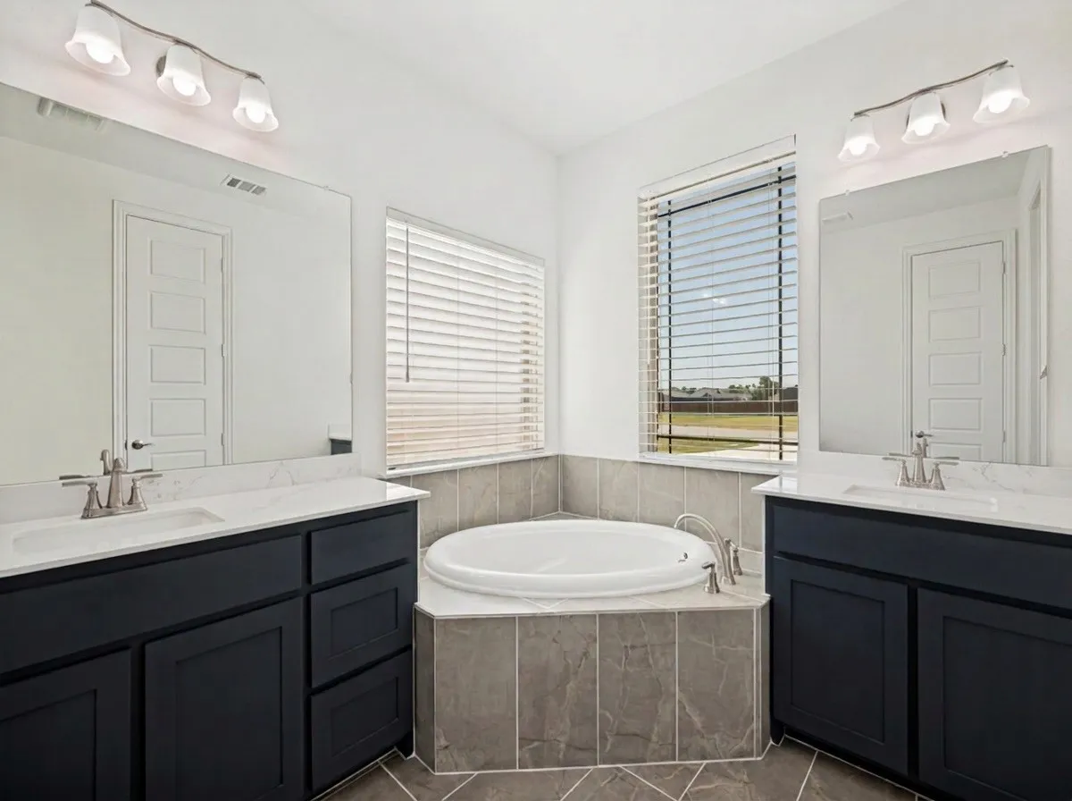 Primary Bathroom featuring dual vanities with black cabinetry and marble countertops, large framed mirrors with integrated lighting, soaking tub with tile surround, and dual window treatments providing natural light and privacy