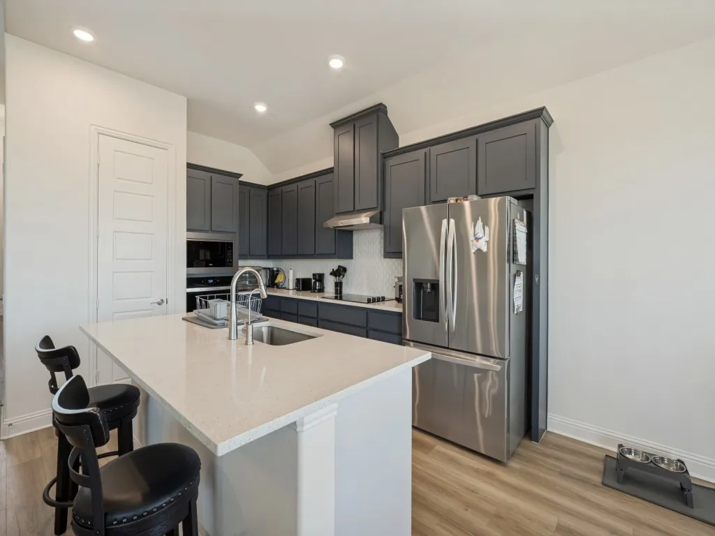 Modern kitchen featuring dark cabinetry, stainless steel appliances, a white quartz countertop island with an undermount sink, and wood-finish flooring