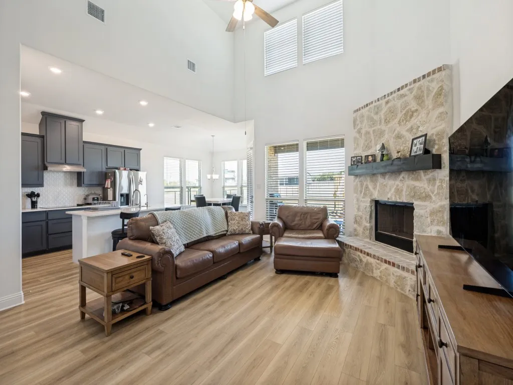 Vaulted ceiling with ceiling fan, stone fireplace with hearth, wood-finish flooring, white shaker island, and dark cabinetry