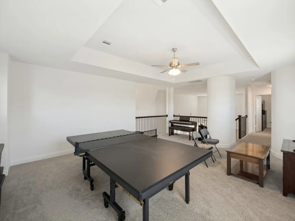 Recessed tray ceiling with a built-in ceiling fan, light gray carpet flooring, and white painted walls