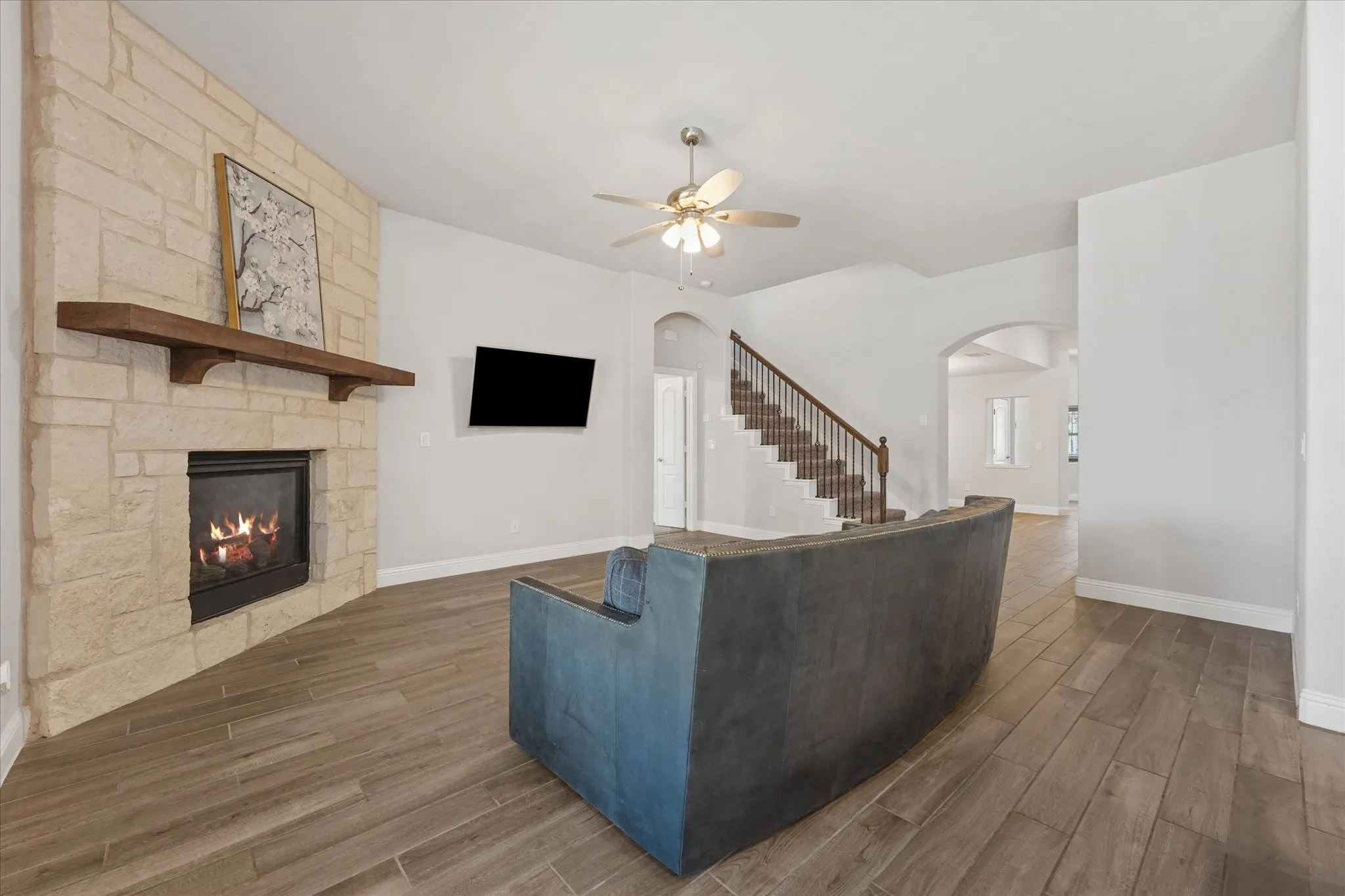 Stone-clad fireplace with a wood mantel, wood-finish flooring, staircase with wood handrails, and an arched entryway