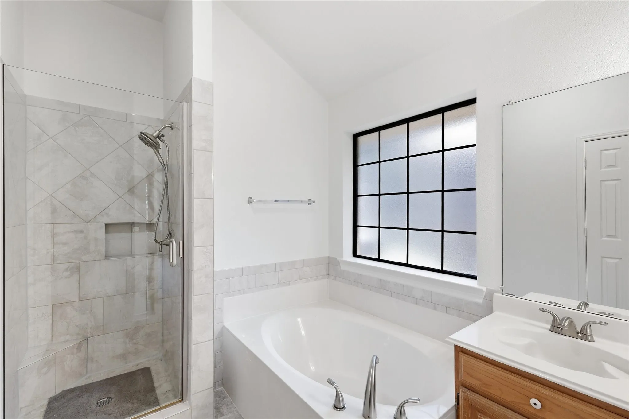 Enclosed shower with tiled walls and built-in niche, soaking tub with tile surround, wood-finish vanity with integrated sink, and privacy window with grid