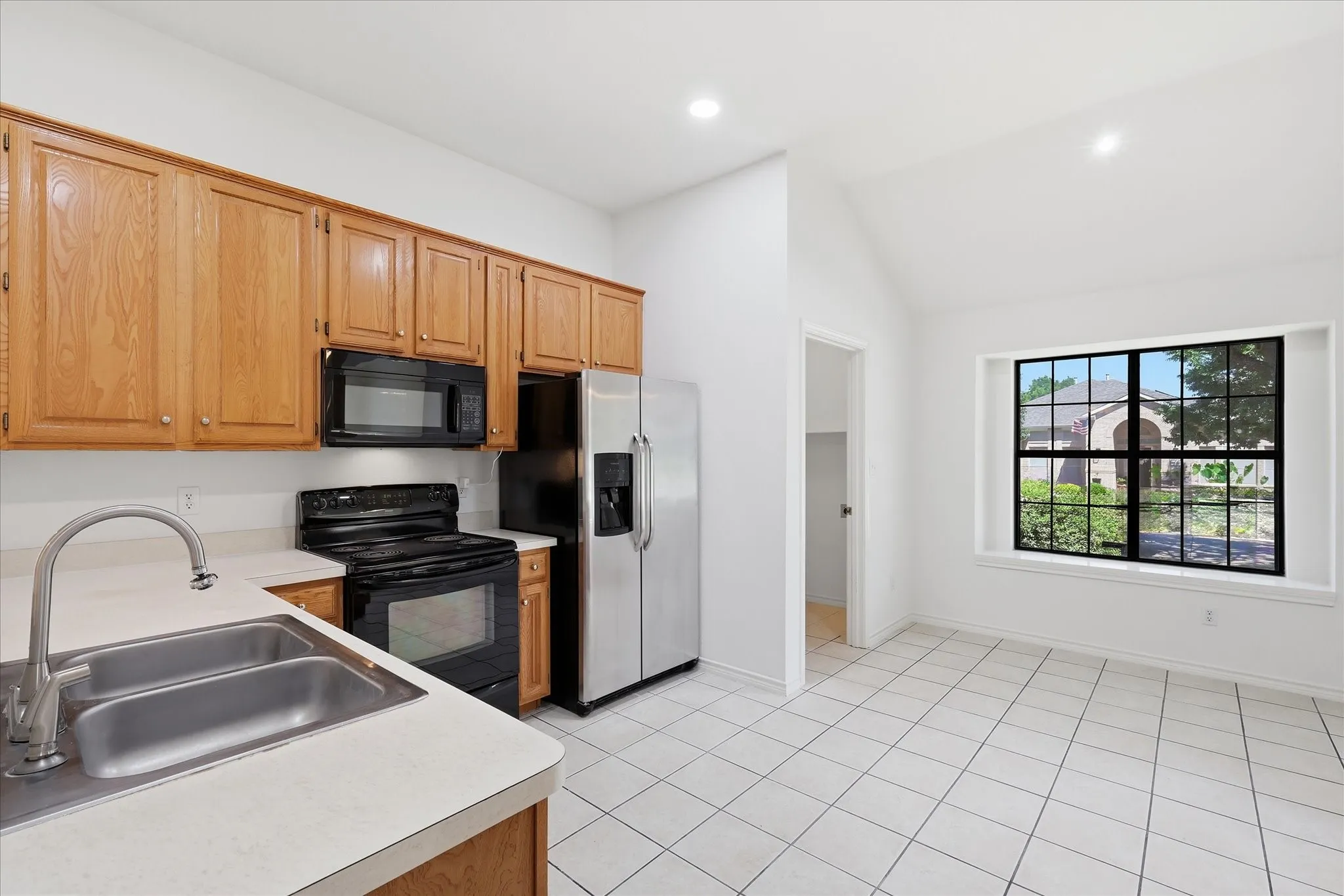 Spacious kitchen featuring warm wood-finish cabinetry, stainless steel appliances, white tile flooring, and a large window framing natural light and outdoor scenery