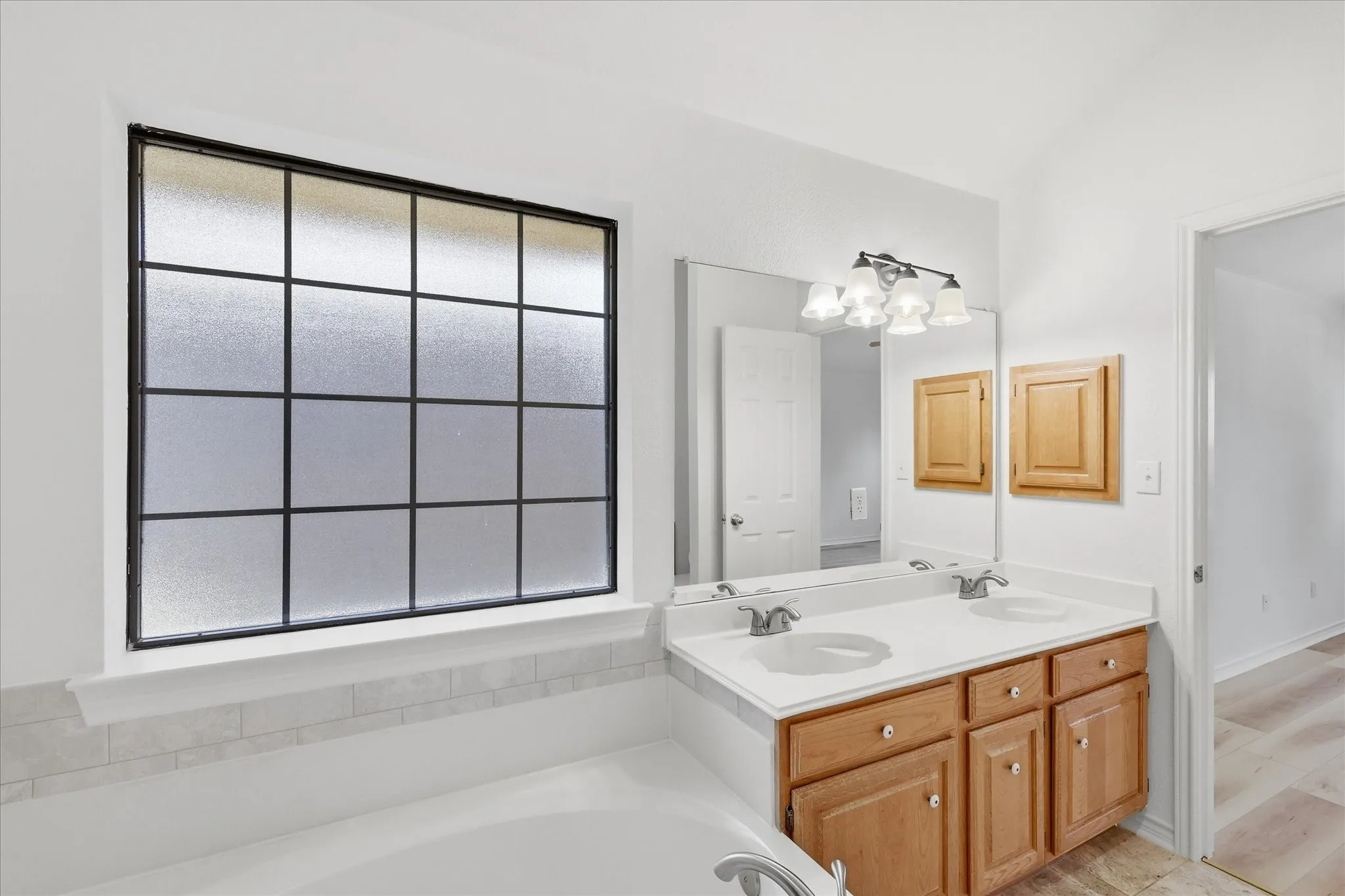 Frosted grid-pattern window above a built-in tub, dual sink vanity with integrated countertop, and warm wood-finish cabinetry