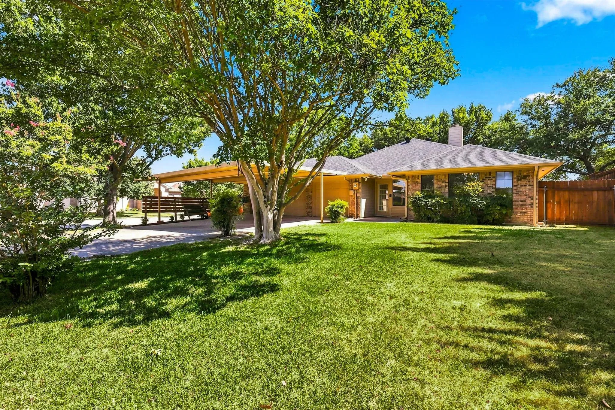 Brick exterior home with a gabled roof, attached carport, mature trees, and a spacious front lawn
