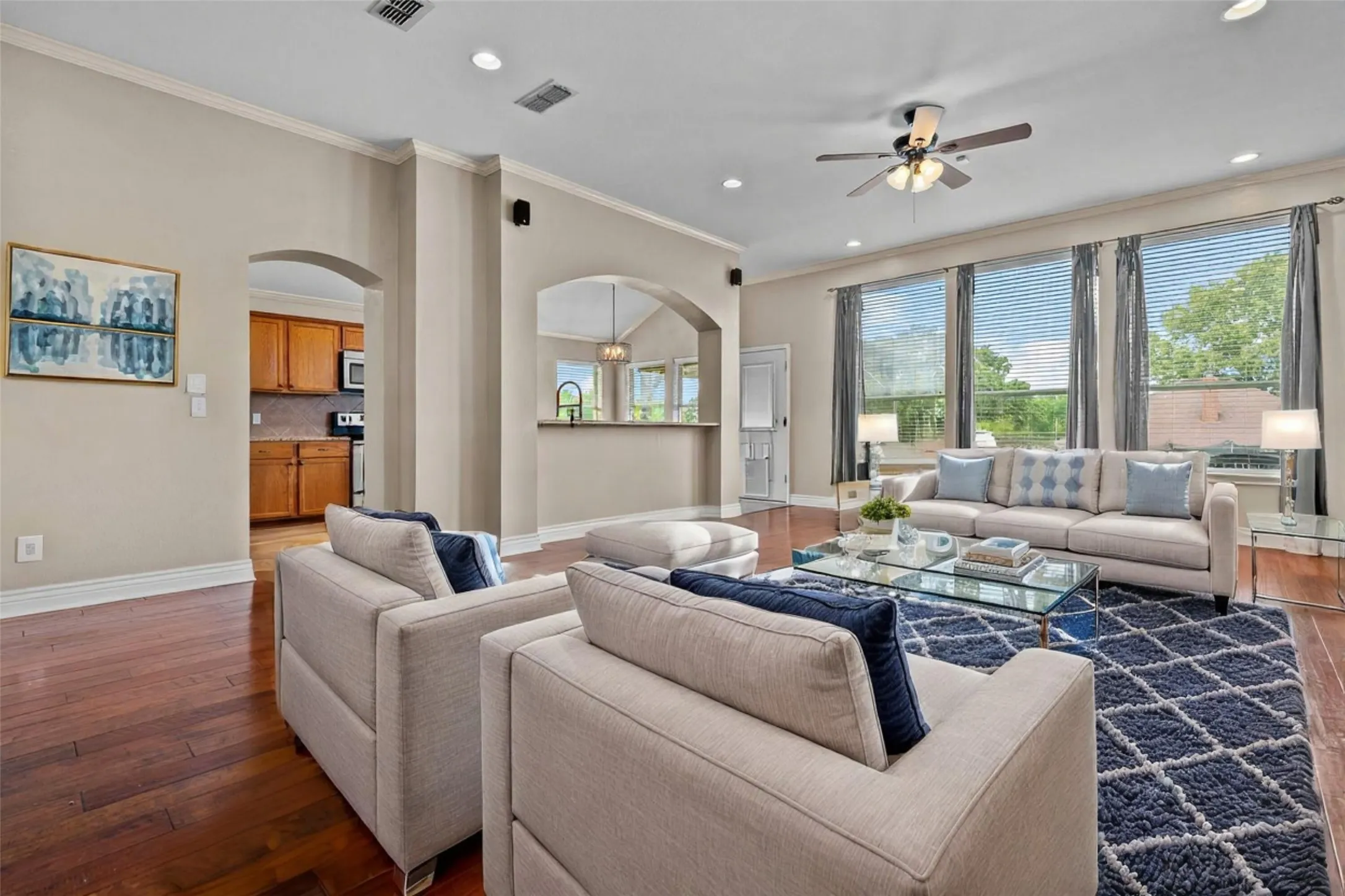 Spacious open-concept living area featuring hardwood flooring, multiple large windows with natural light, architectural columns, ceiling fan with recessed lighting, neutral wall finishes, and a gas fireplace with decorative surround
