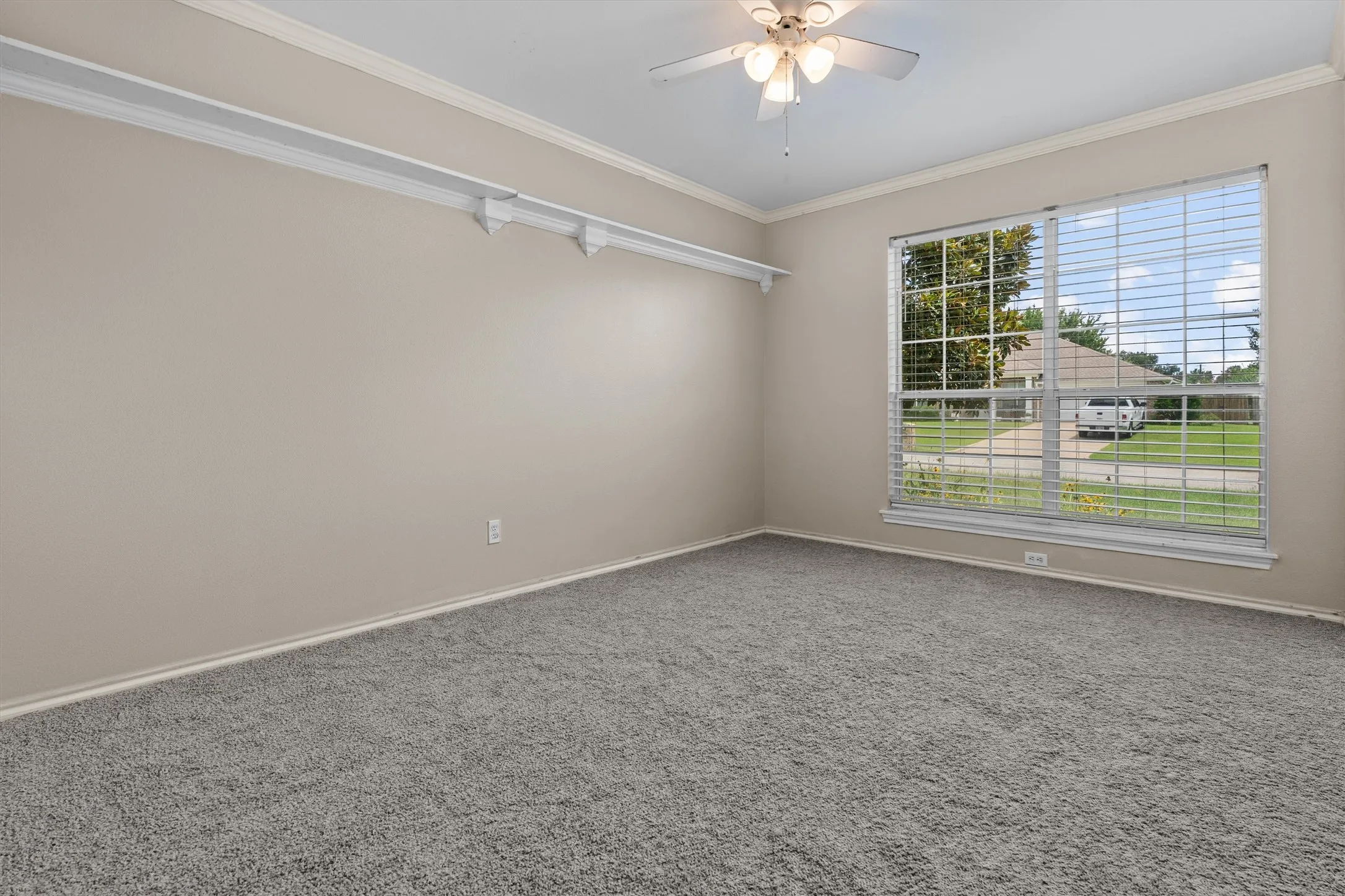Spacious room showcasing neutral wall finishes, plush carpeting, abundant natural light from large windows with plantation shutters, ceiling fan with integrated lighting, and track lighting along the ceiling for flexible illumination
