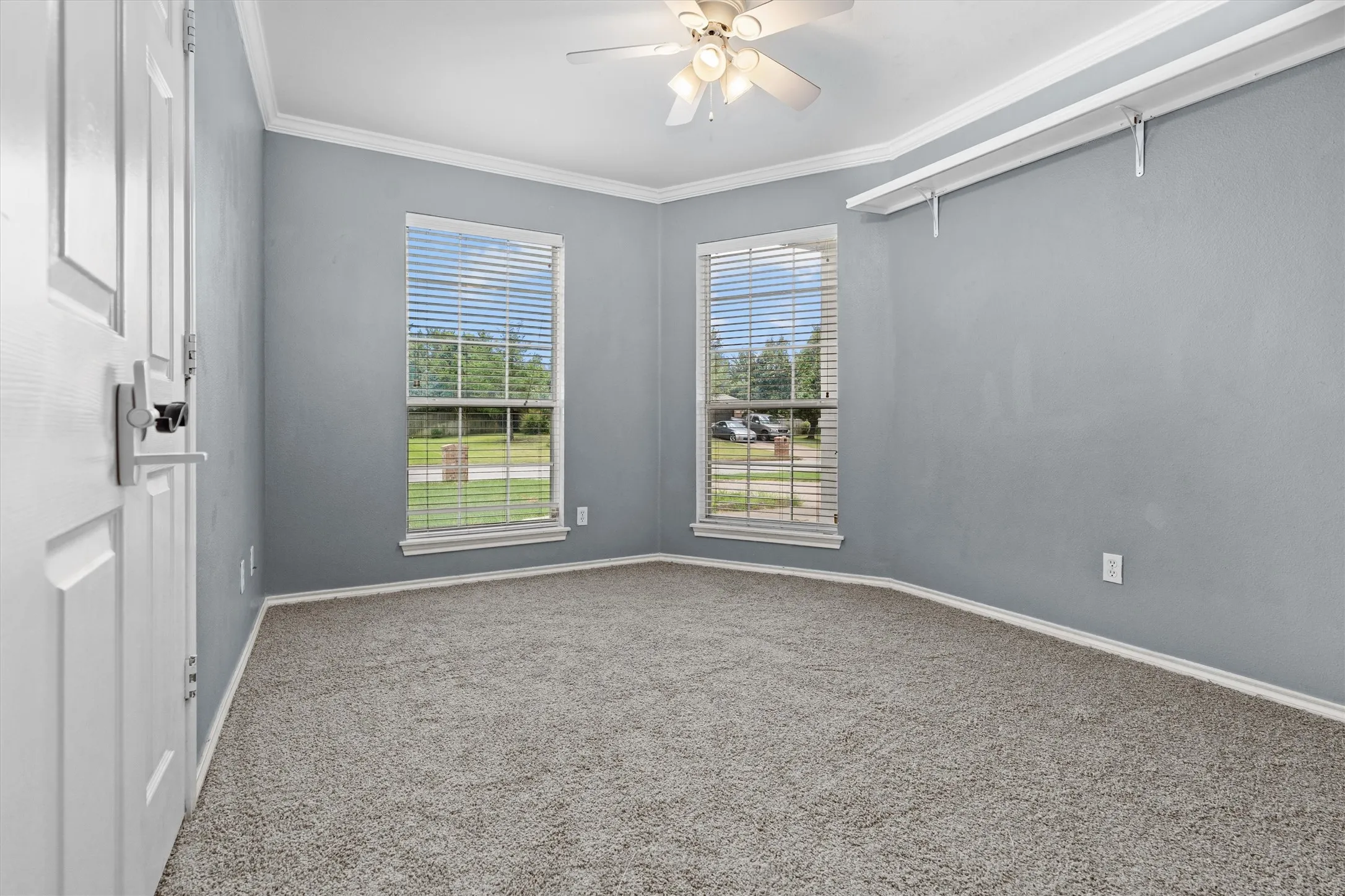 Bright corner bedroom with dual windows framing outdoor scenery, neutral gray walls, neutral carpet flooring, and ceiling fan with integrated lighting