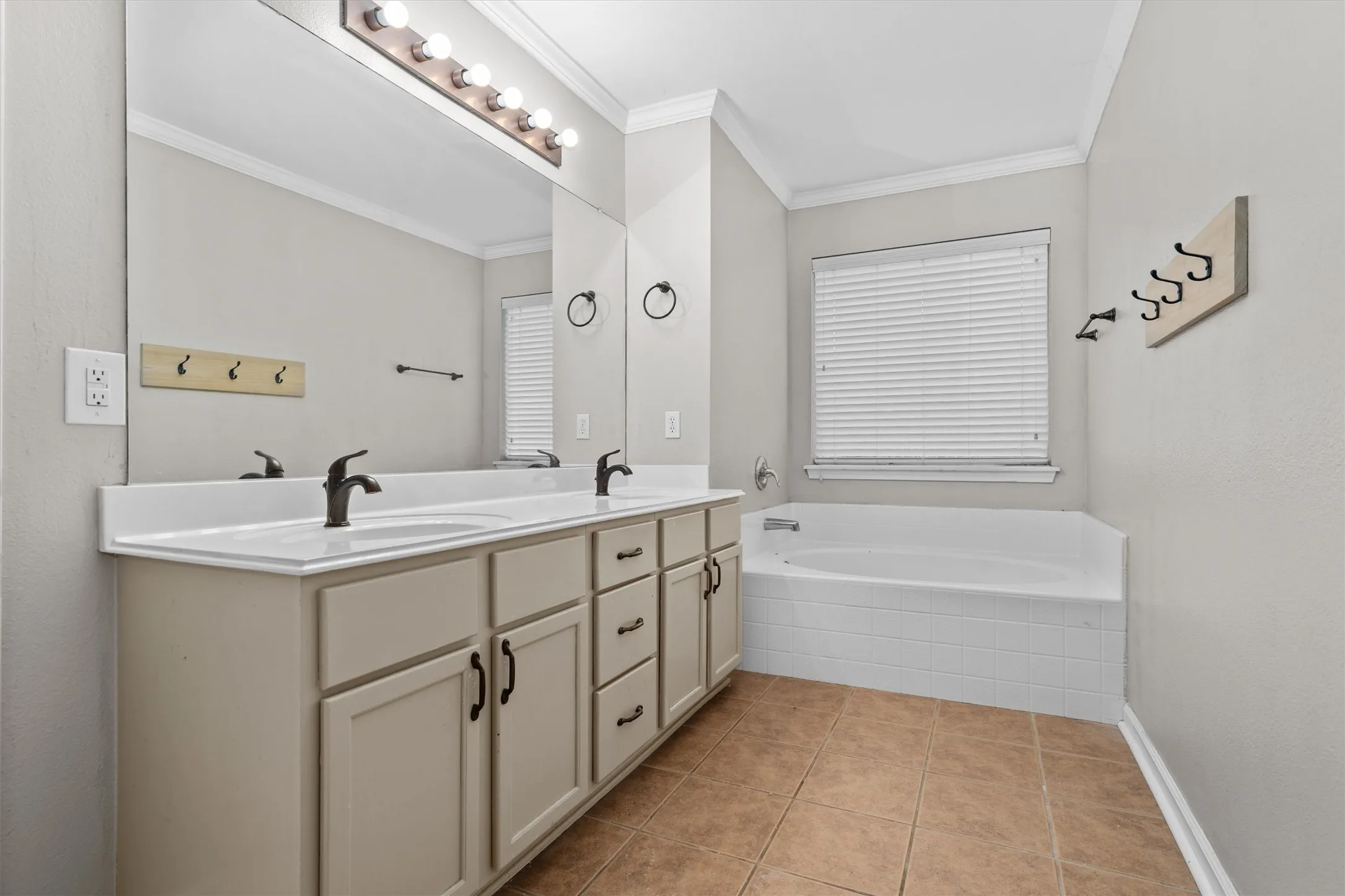 Primary bathroom featuring a dual-sink vanity with wood-finish cabinetry, white countertop, and matte black faucets; framed mirror with integrated lighting; soaking tub with tile surround; tile flooring; and a window with plantation shutters