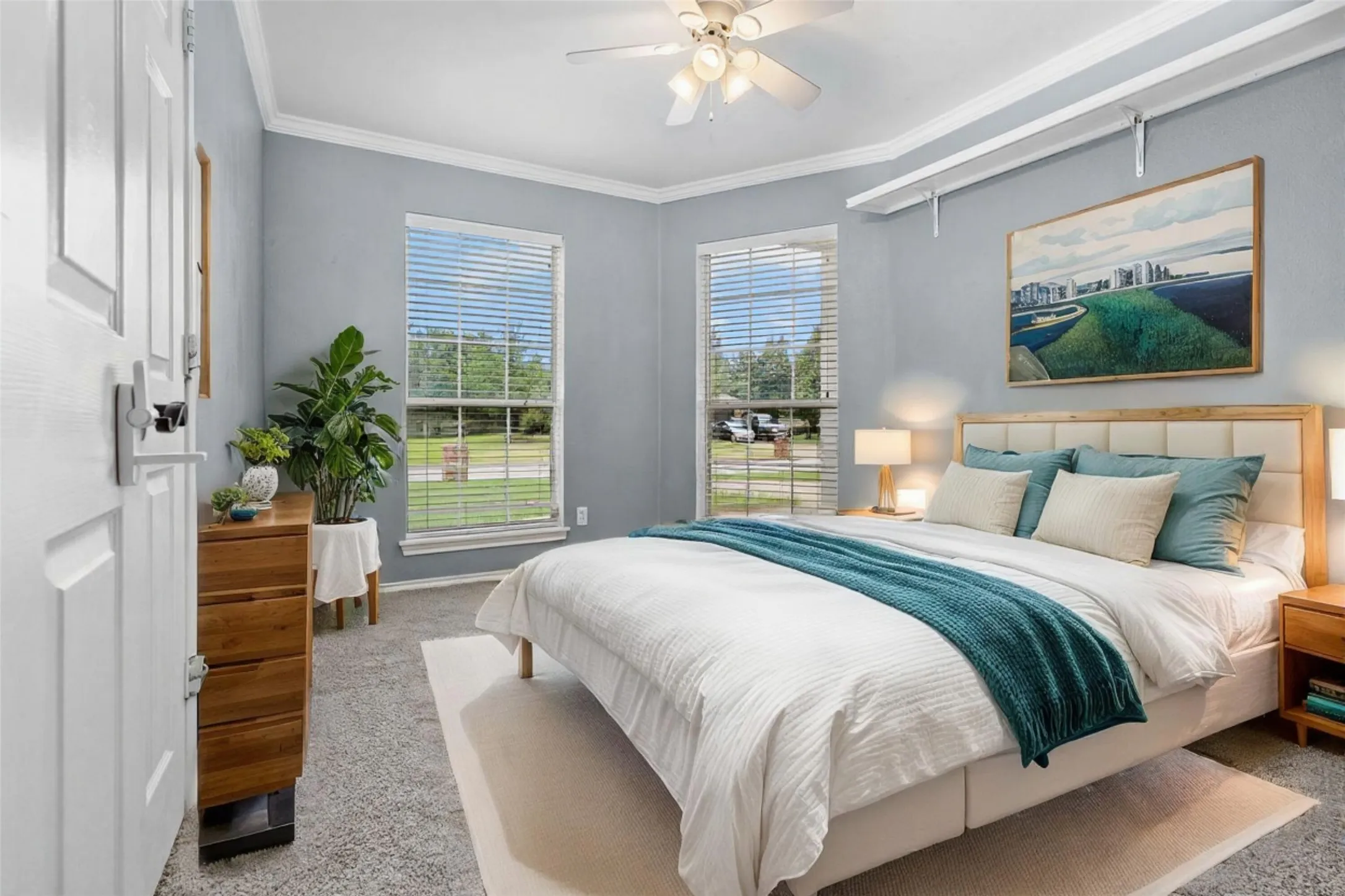 Serene primary bedroom suite featuring dual glass doors opening to outdoor landscape, soft blue-gray wall finishes, wood-finish nightstands with integrated lighting, ceiling fan with light fixture, and plush bedding in complementary tones