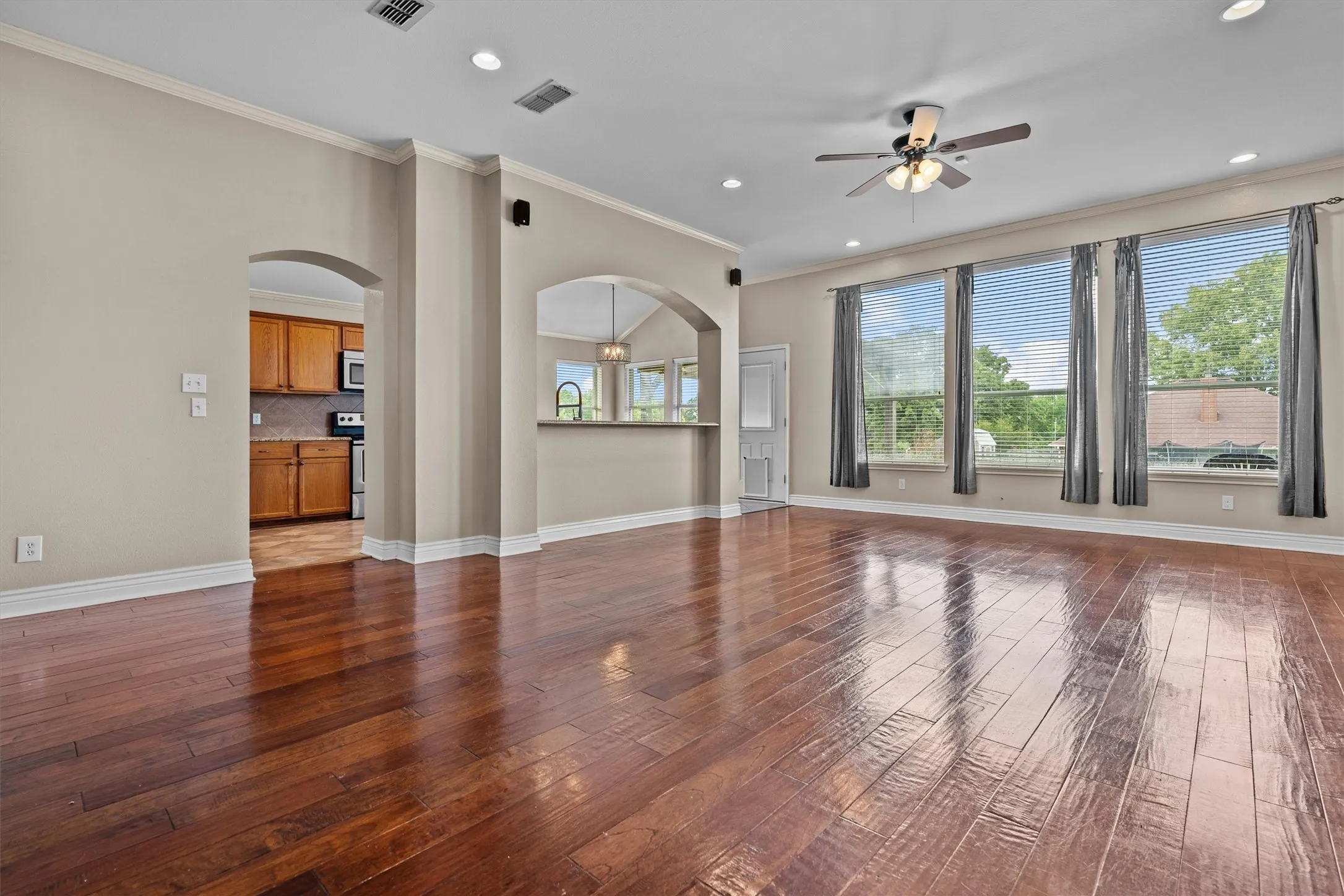 Expansive open-concept living area showcasing rich wood-finish flooring throughout, soaring ceilings with recessed lighting and ceiling fan, and abundant natural light from multiple large multi-pane glass doors and windows