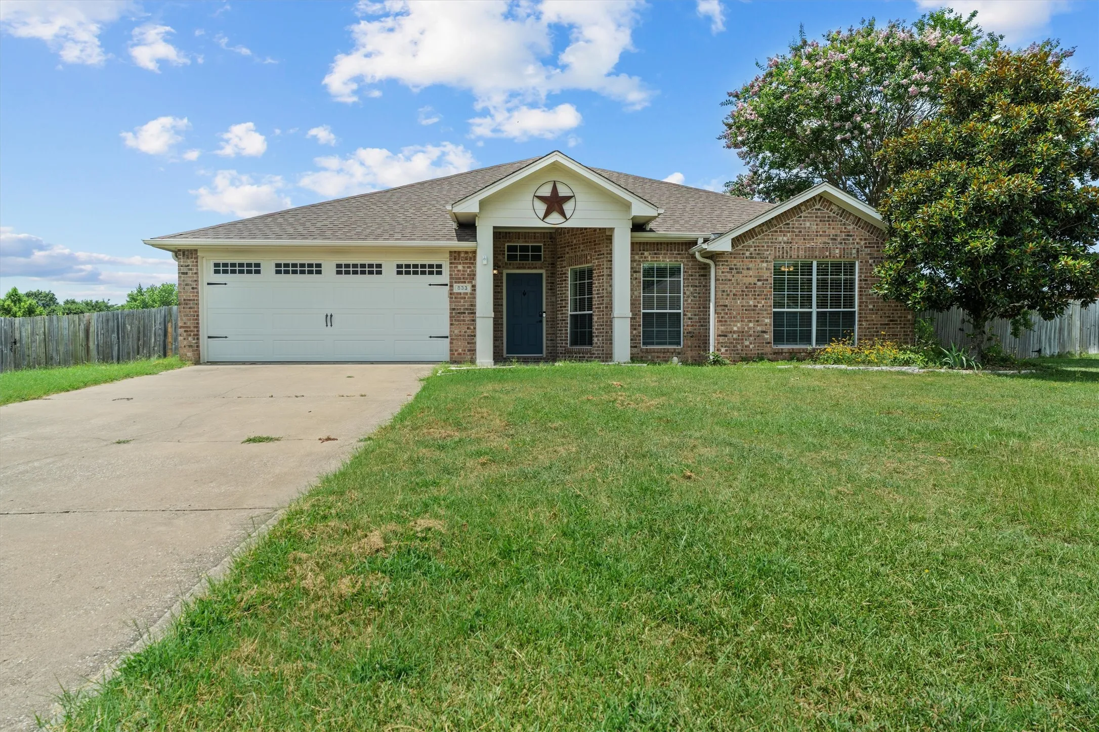 Charming one-story brick residence featuring a two-car garage with paneled doors, arched entryway with decorative star accent, multiple large windows with black trim, and a spacious front lawn