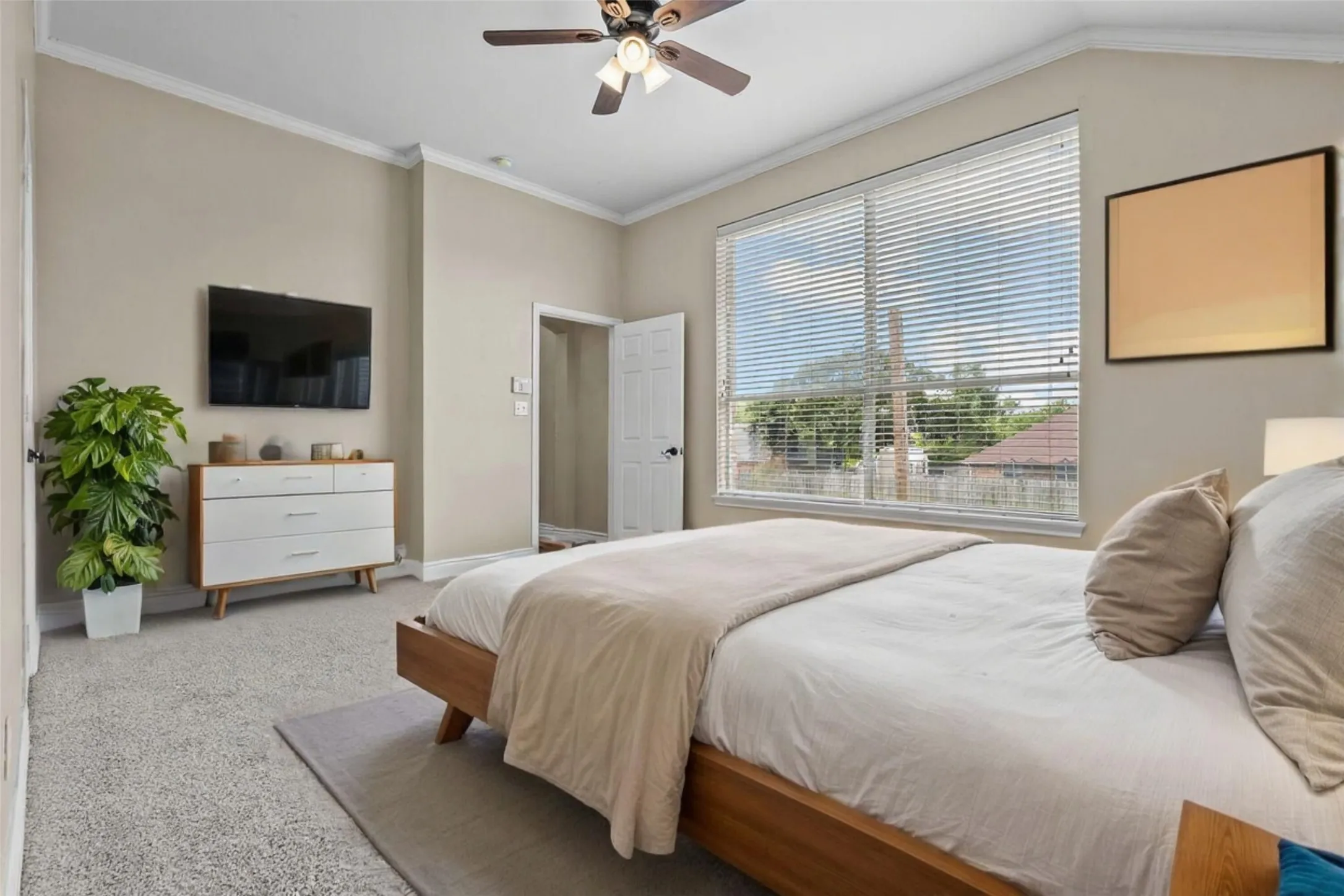 Spacious primary bedroom featuring a wood-finish platform bed, soft neutral palette, ceiling fan with integrated lighting, wall-mounted flat-screen television, built-in white cabinetry with decorative shelving, and expansive sliding glass doors with horizontal blinds opening to natural light and outdoor views