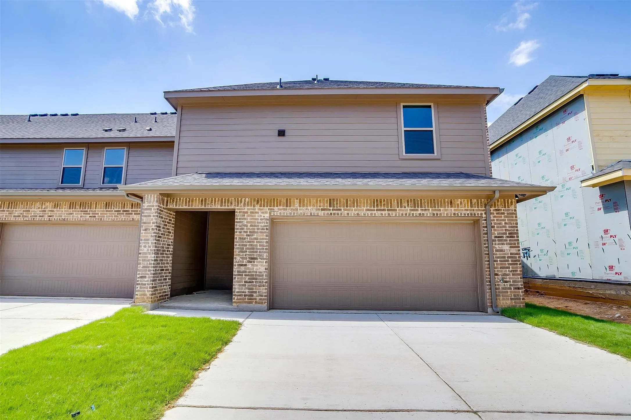 Two-car garage with neutral-toned paneled doors, brick and siding exterior with contrasting trim, manicured lawn, concrete driveway, and contemporary two-story design featuring multiple windows and clean architectural lines