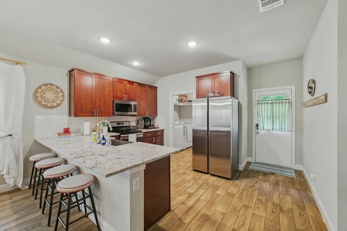 Kitchen featuring wood-finish flooring, cherry-toned cabinetry, stainless steel appliances, a white tile backsplash, and a light-toned countertop with a breakfast bar