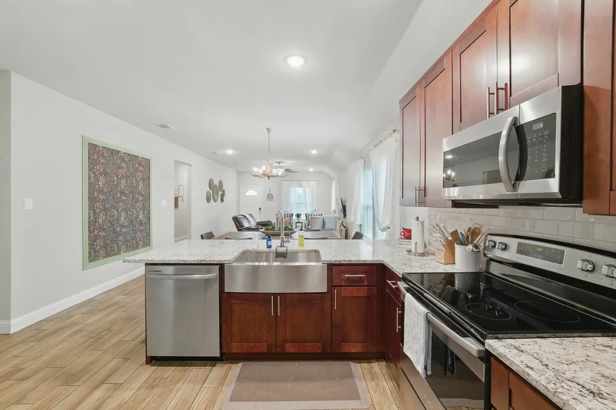 Open-concept kitchen featuring wood cabinetry, stainless steel appliances, a farmhouse sink, stone countertops, and a subway tile backsplash