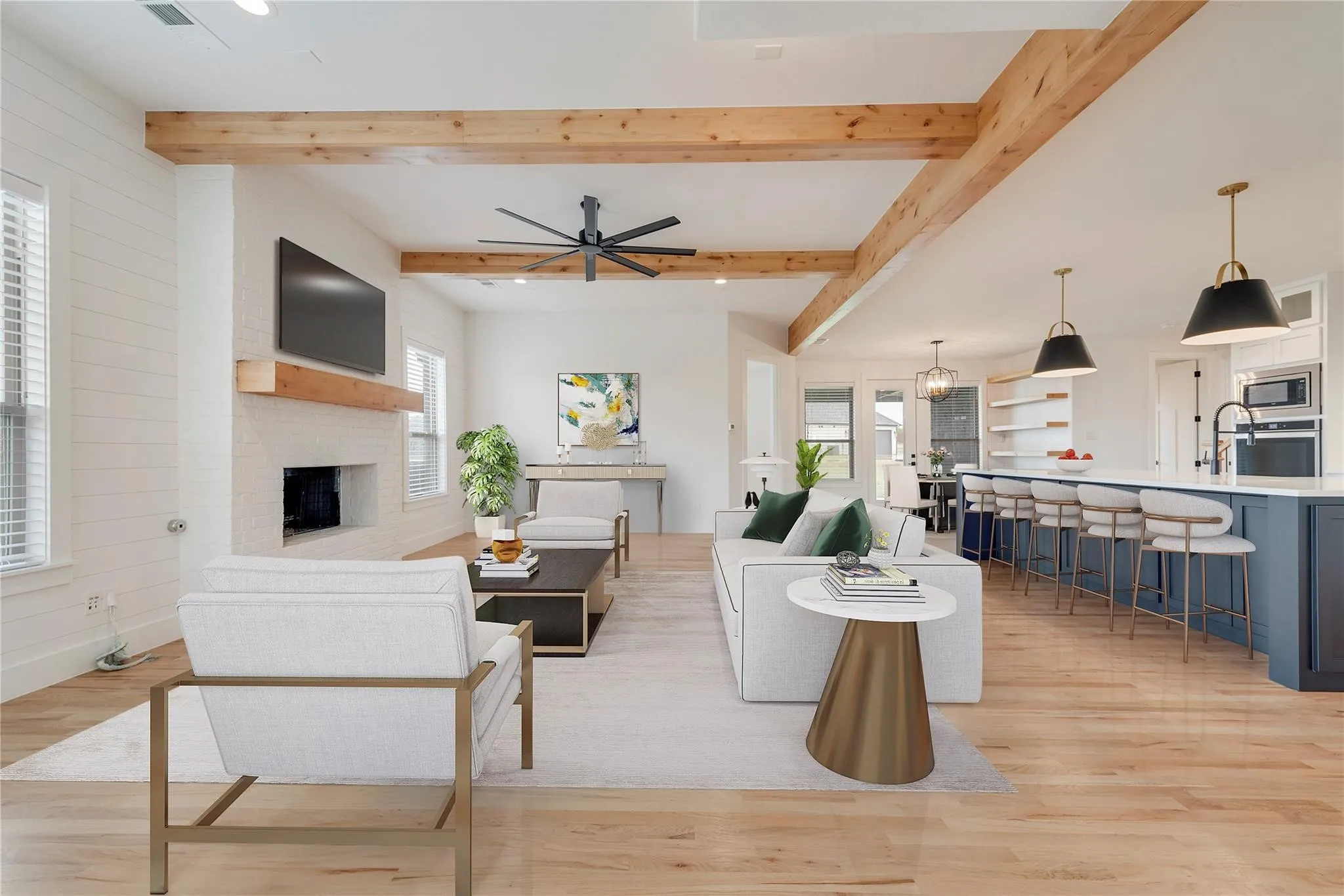 Spacious open-concept living area featuring exposed wood ceiling beams, wood-finish flooring, a white brick fireplace with a wood mantel, and large windows