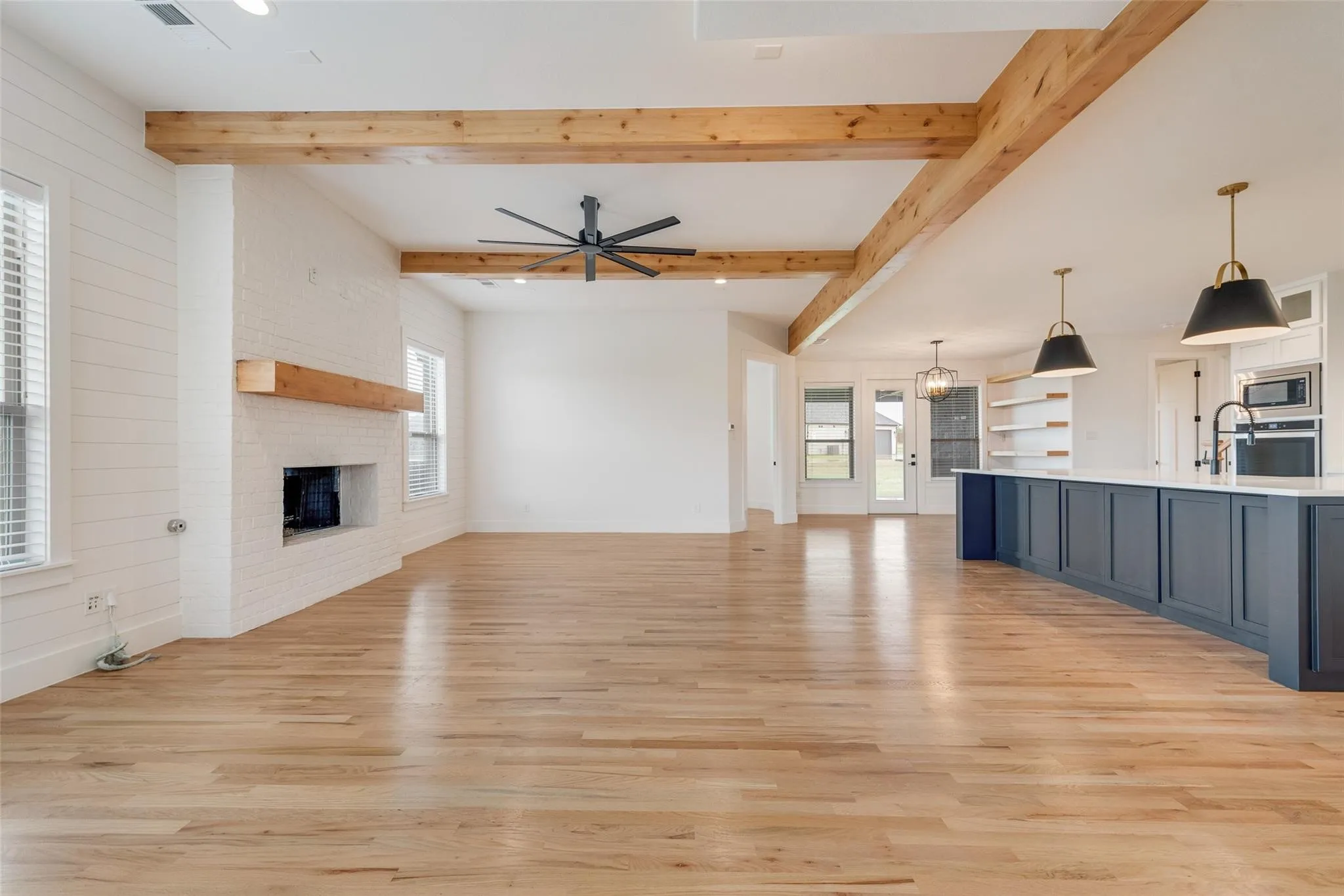 Expansive open-concept living area featuring blonde wood-finish flooring, exposed wood beams, and a white brick fireplace with a wood mantel
