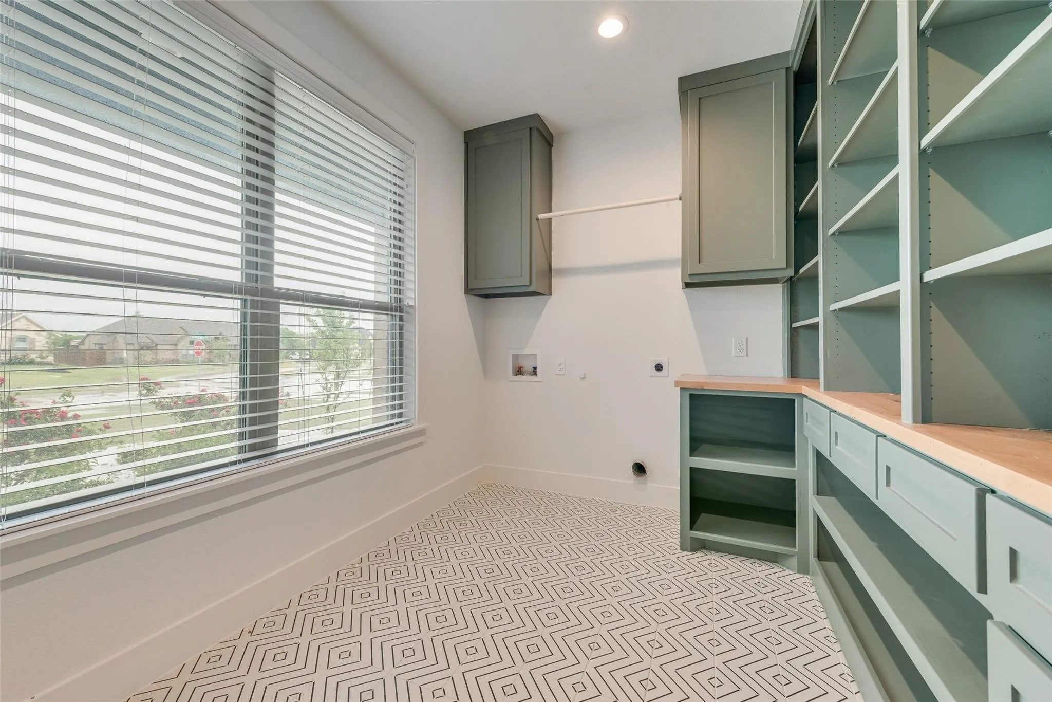 Laundry room featuring patterned tile flooring, custom green cabinetry, built-in shelving, wood-finish countertops, and a large window with blinds