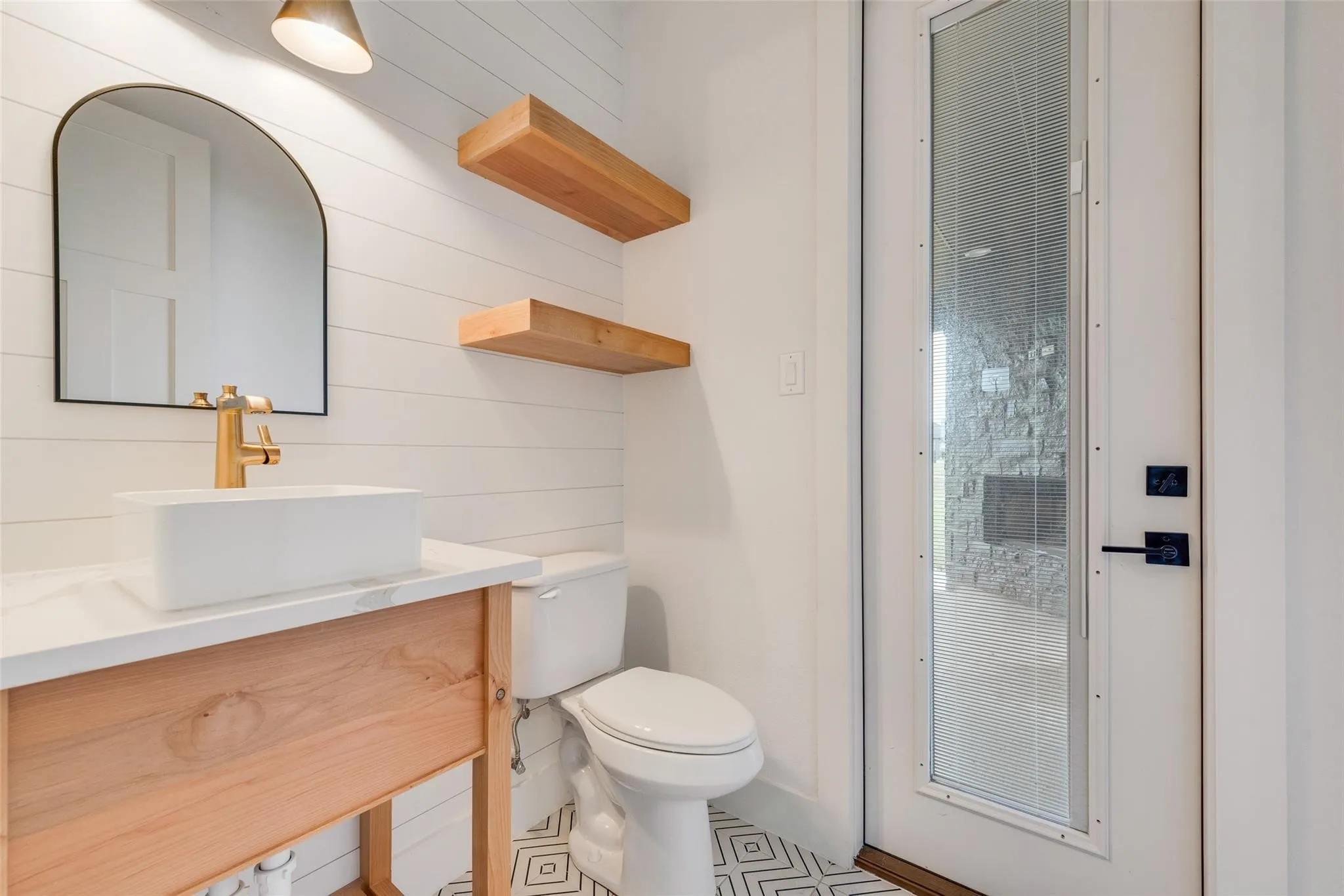 Powder room featuring shiplap wall paneling, a wood-finish vanity with a vessel sink and gold-tone faucet, a black-framed arched mirror, and open shelving