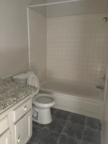 White tile surround bathtub with shower fixture, dark tile flooring, white vanity with a speckled countertop, and a white toilet