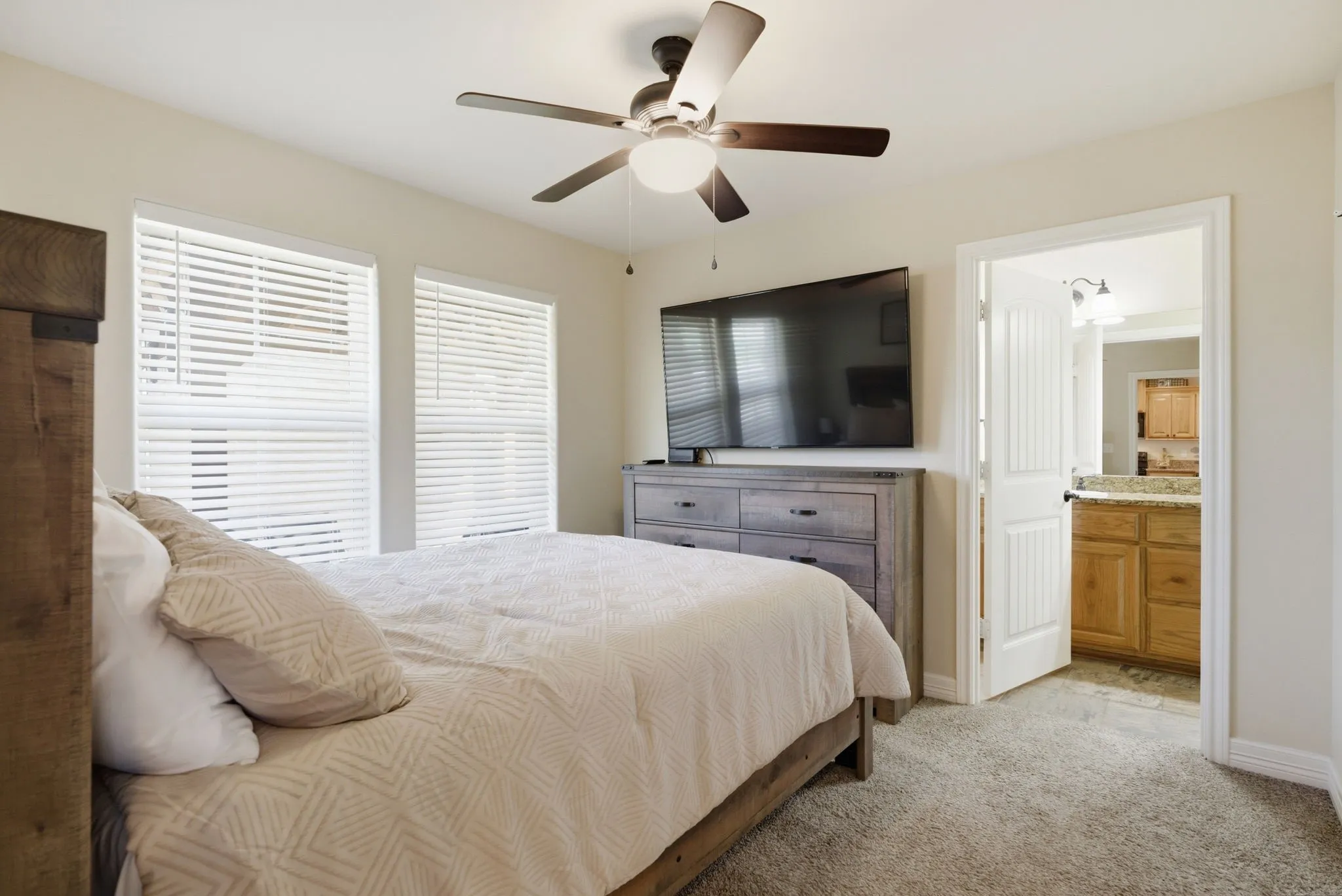 Carpeted bedroom with a ceiling fan, two windows with blinds, and an open doorway revealing a bathroom with wood cabinetry and a stone countertop