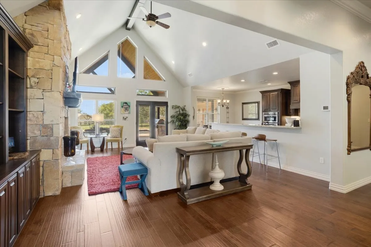 Vaulted ceiling with exposed beam, stone-clad fireplace, wood-finish flooring, built-in cabinetry, and a kitchen pass-through with a breakfast bar