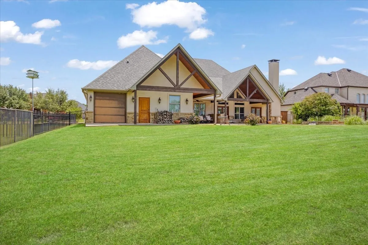 Expansive green lawn with a black metal fence, covered patio featuring exposed wood beams, and a stucco and stone exterior