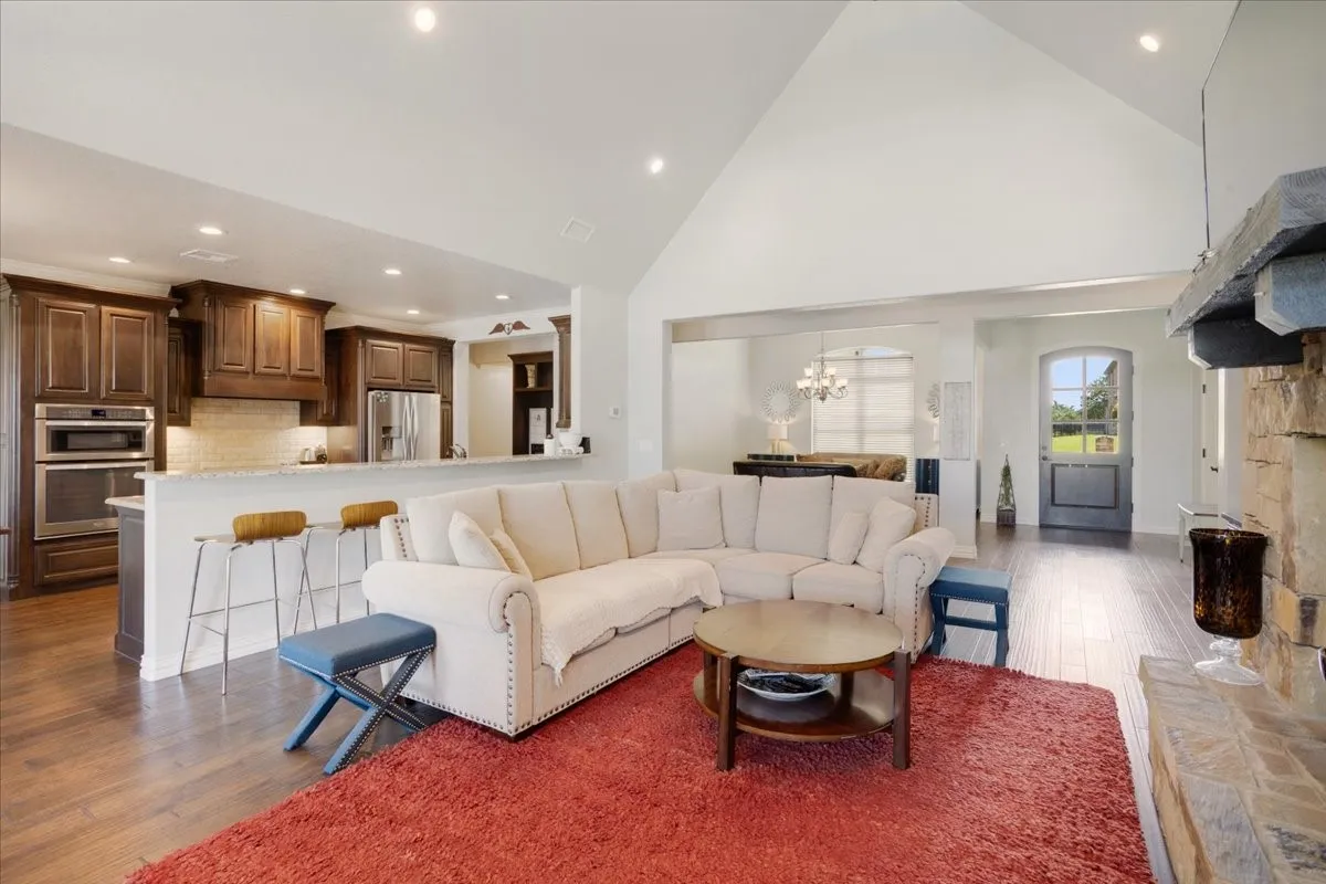 Spacious open-concept living area with vaulted ceilings, wood-finish flooring, a stone fireplace, and a kitchen featuring dark wood cabinetry and stainless steel appliances