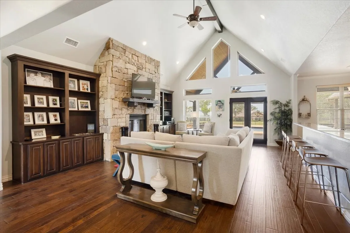 Vaulted ceiling with exposed beams, stone-clad fireplace, built-in cabinetry, wood-finish flooring, and a breakfast bar