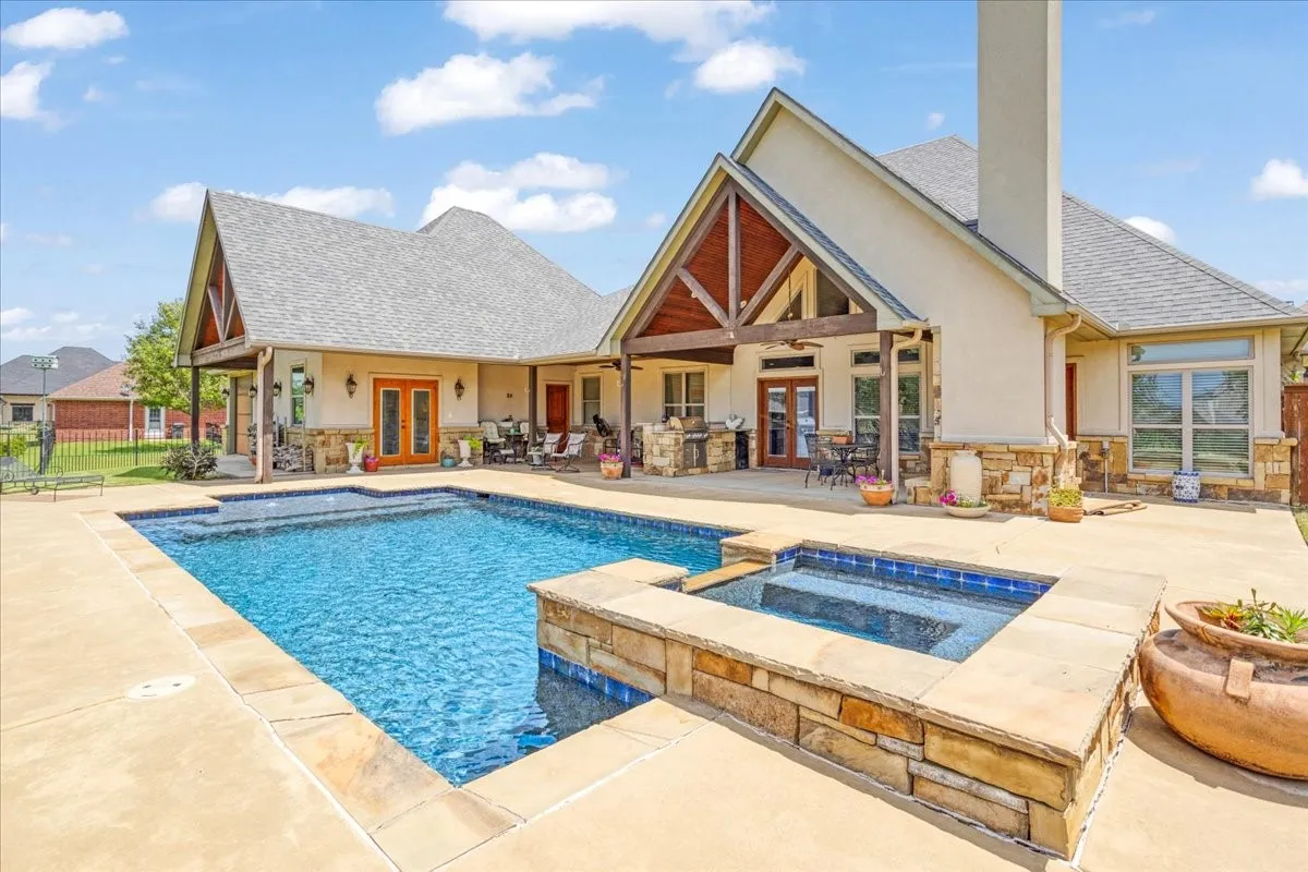 In-ground swimming pool with integrated spa, extensive concrete decking, and covered patio featuring exposed wood beams and stone accents
