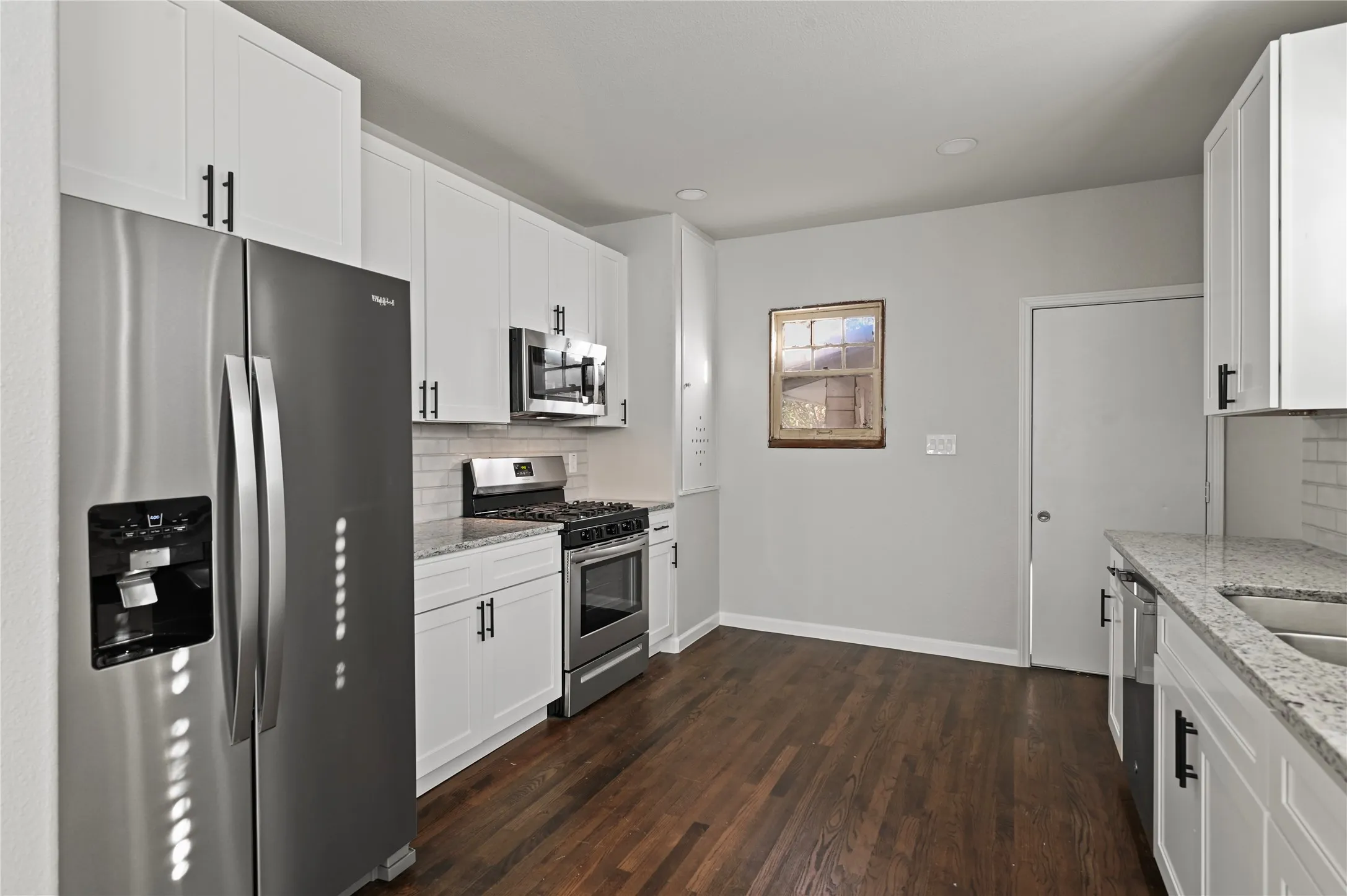 White cabinetry with matte black hardware, stainless steel appliances, speckled stone countertops, subway tile backsplash, and dark wood-finish flooring