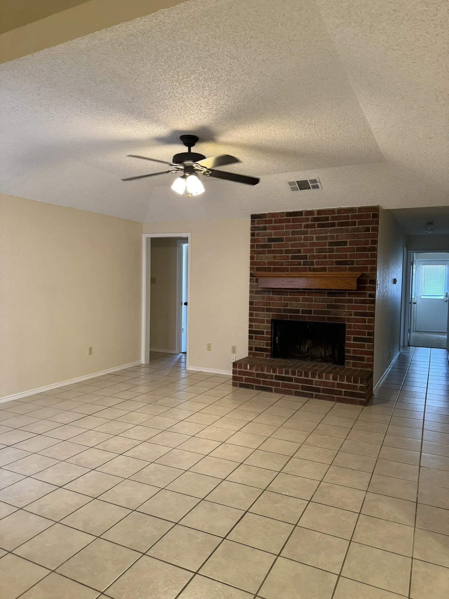 Brick-surround fireplace with a wood mantle, ceiling fan with integrated lighting, and light-toned tile flooring
