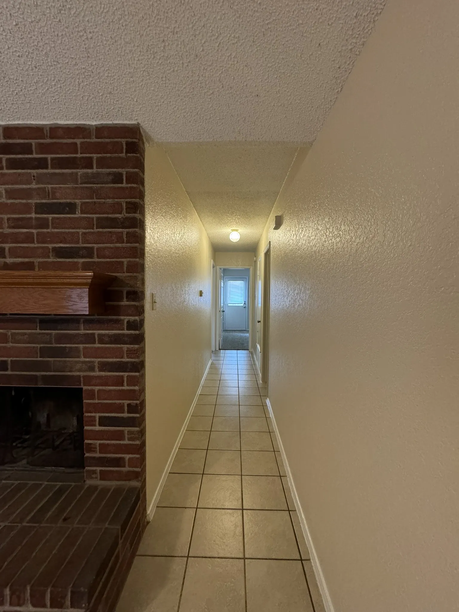 Brick fireplace with wood mantel, tile flooring, textured walls, and overhead lighting