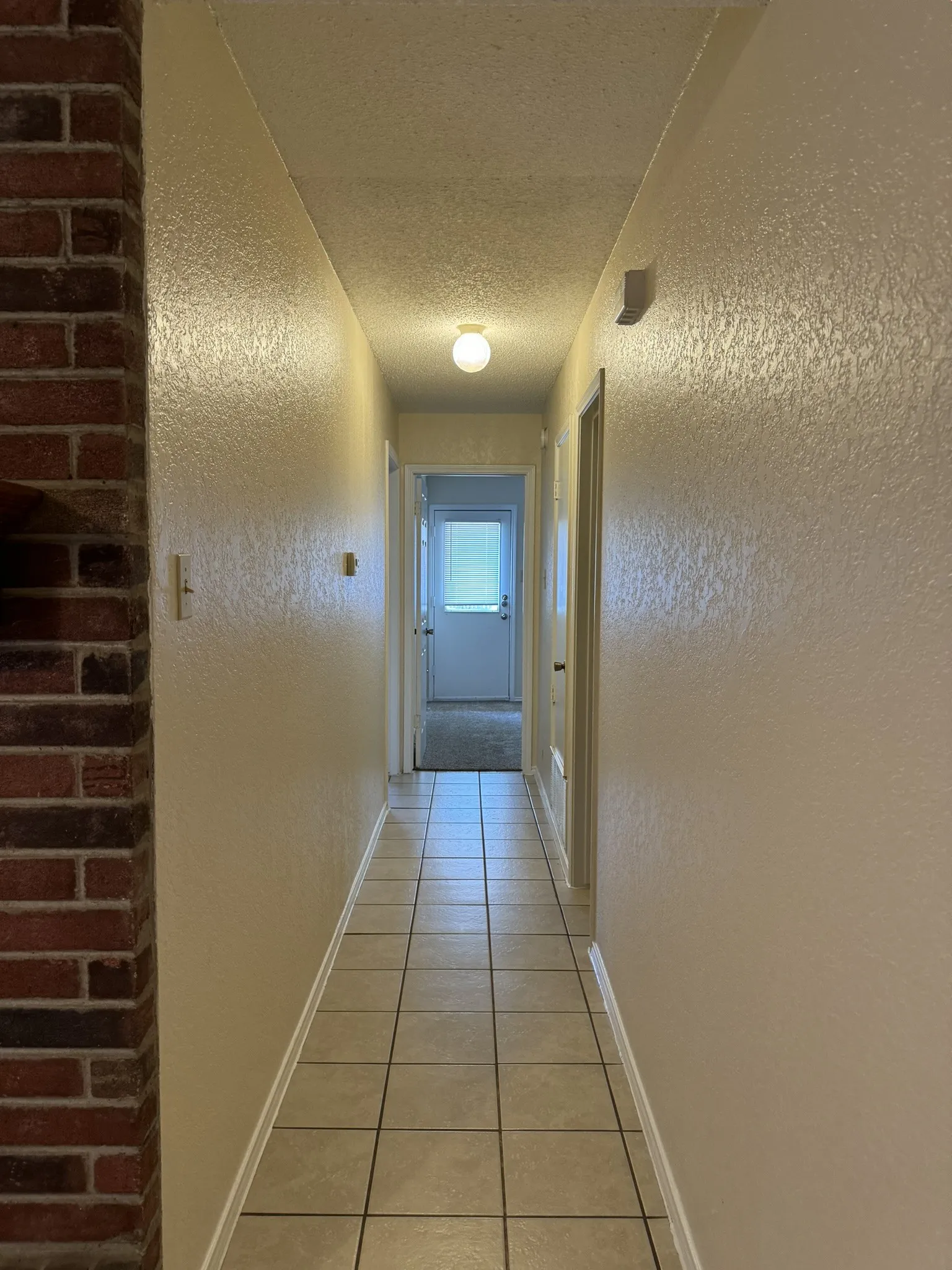 Hallway featuring tile flooring, textured walls, a ceiling-mounted light fixture, and a brick-faced architectural element