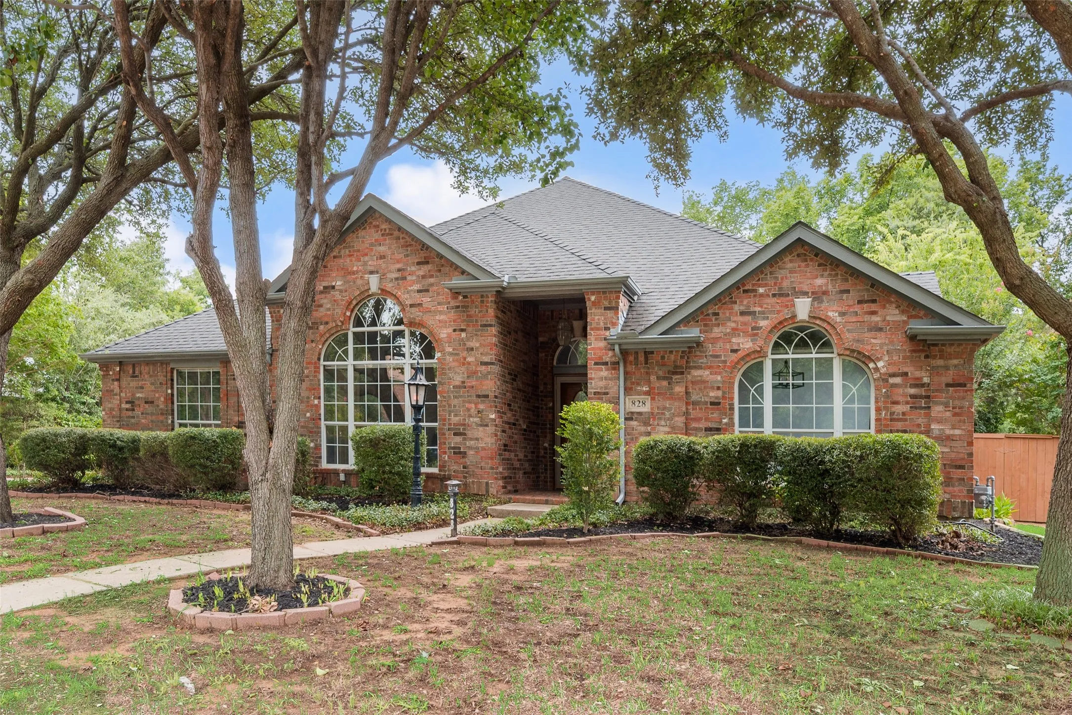 Brick exterior with a gray shingle roof, arched windows, and established landscaping