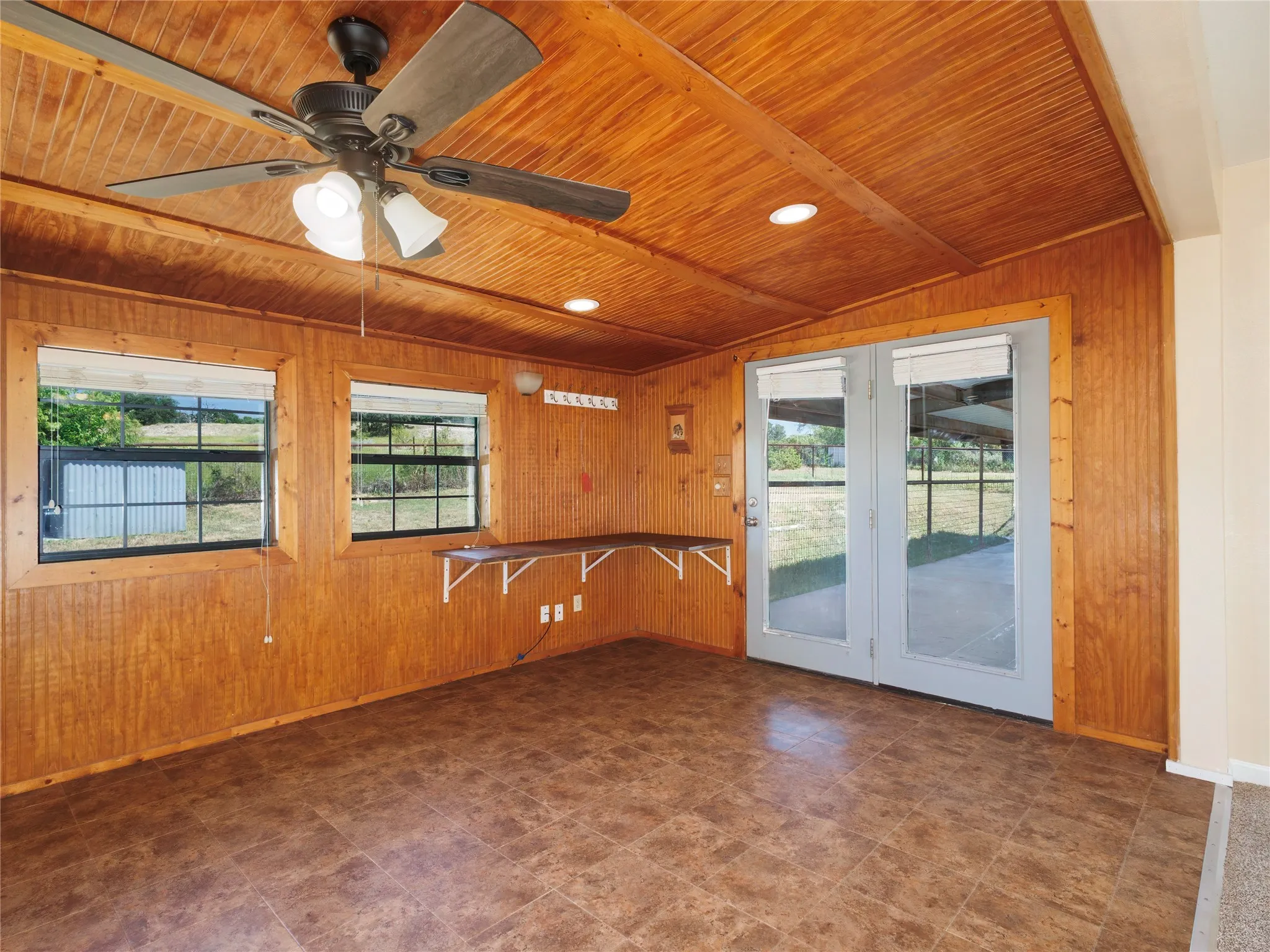 Wood-paneled walls and ceiling, two large windows, a built-in shelf, double glass-paneled doors, and a ceiling fan with integrated lighting